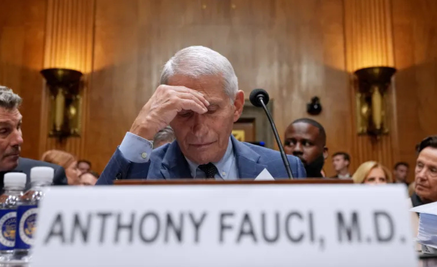 Anthony Fauci, seated at a Senate hearing table with a nameplate reading "Anthony Fauci, M.D.," presses his hand to his forehead with his eyes closed.
