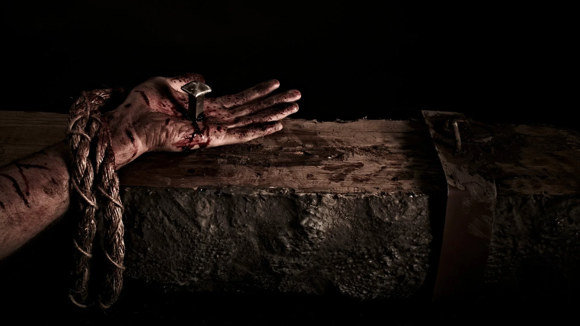 A wounded hand bound with rope and pierced by an iron nail rests on a rough wooden beam against a black background.