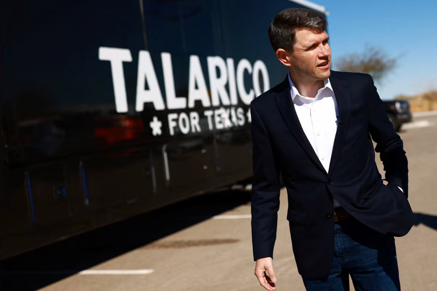 James Talarico, in a dark blazer and jeans, walks past his black “Talarico for Texas” campaign bus in a sunny parking lot.