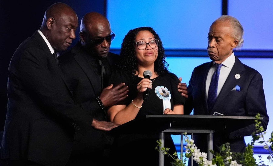 A grieving mother speaks at a podium into a microphone, flanked by her husband and supporters, at a funeral service