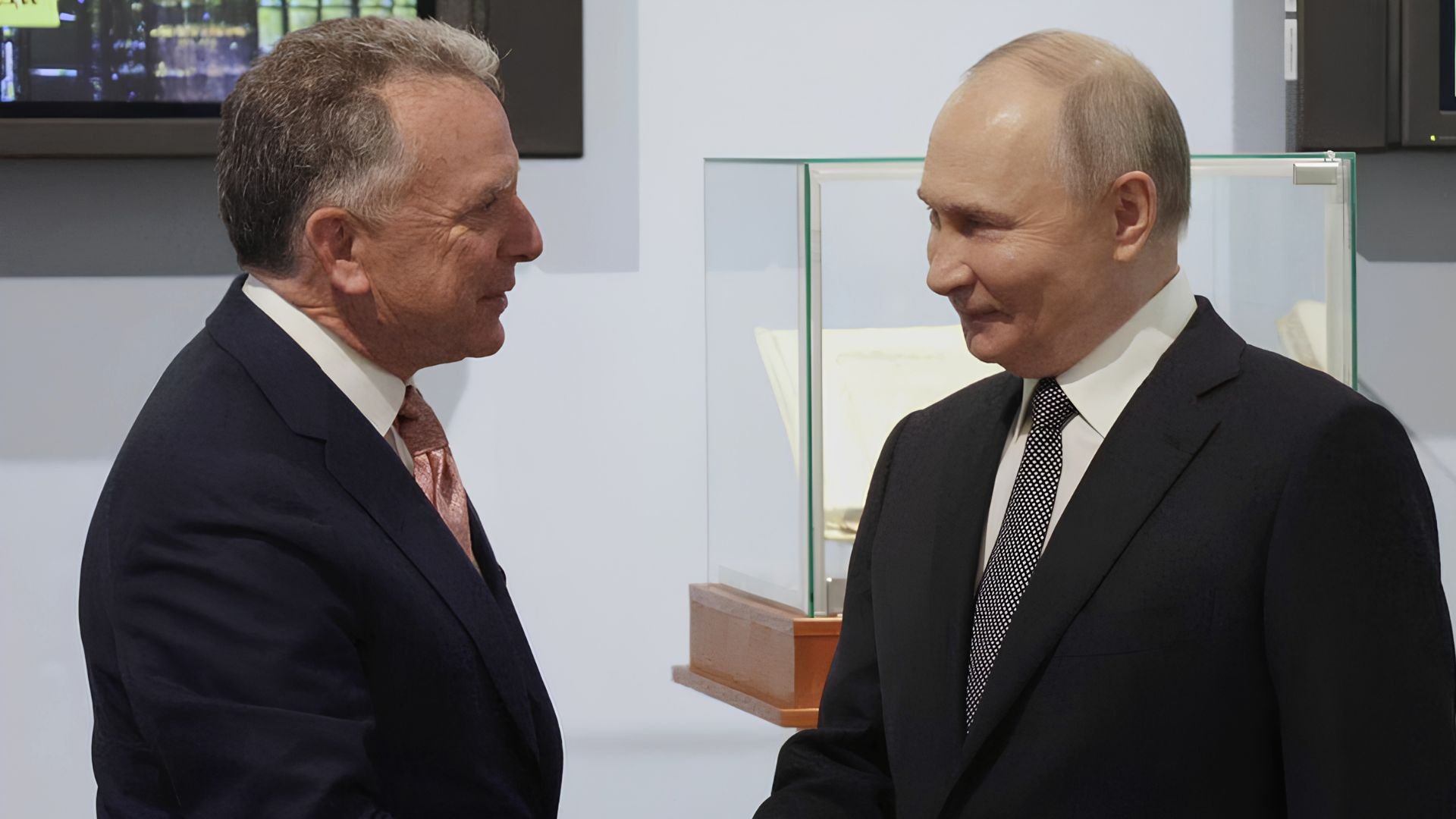 U.S. special envoy Steve Witkoff and Russian President Vladimir Putin, both in dark suits, smile as they shake hands beside a glass display case.