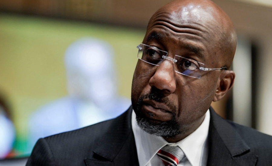 Close-up portrait of Sen. Raphael Warnock wearing glasses, a dark suit, white shirt, and striped tie while looking to the side during an interview.