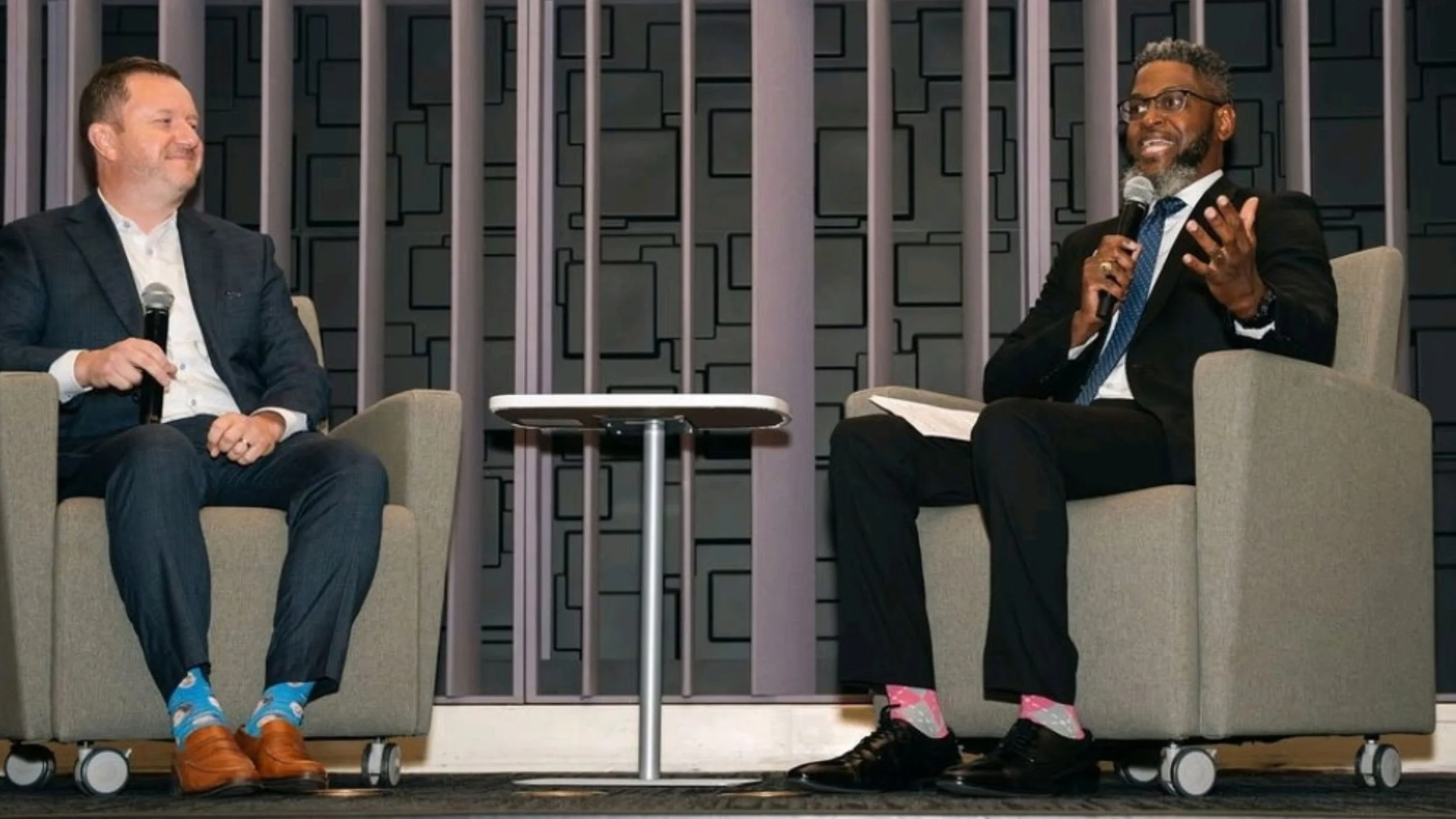 Kenya Hobbs gestures and smiles as he speaks into a microphone from an armchair on stage, while Say Yes Buffalo CEO David Rust listens and smiles from a matching chair beside him.