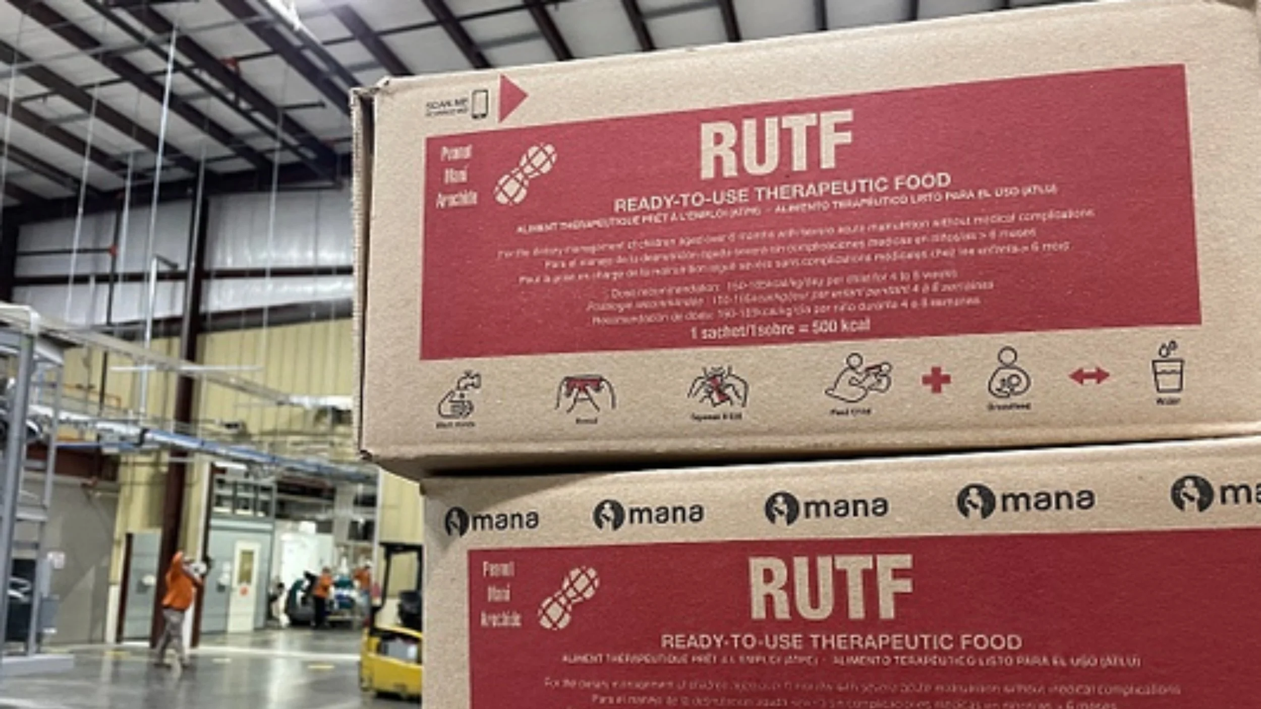 Stacked cardboard boxes printed with “RUTF Ready-to-Use Therapeutic Food” in a factory warehouse, with workers and a forklift in the background.