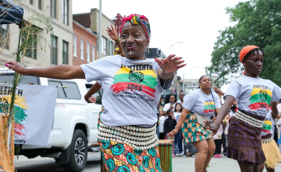 A woman in a colorful headwrap and Juneteenth 1865 T-shirt dances with arms outstretched during an outdoor Juneteenth parade, with other dancers in traditional African-print skirts visible behind her.