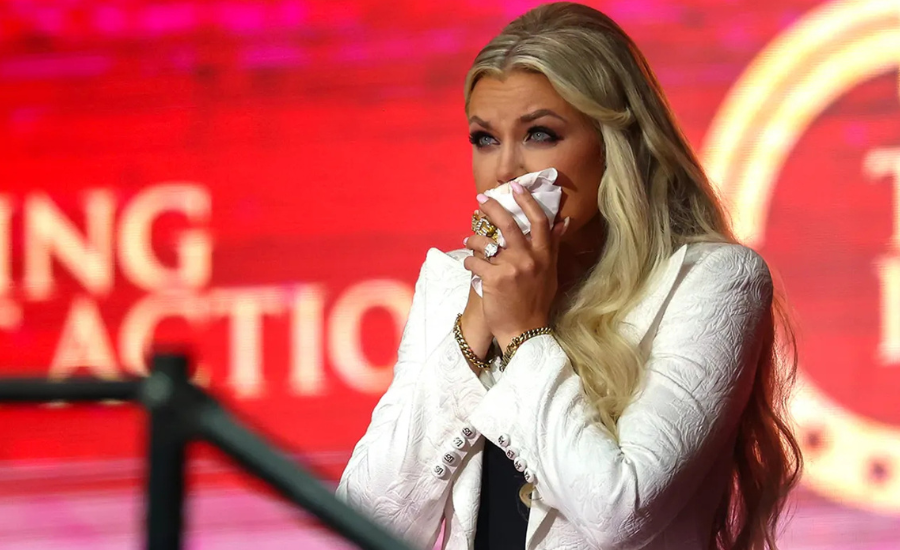 Erika Kirk, wiping tears with a tissue, becomes emotional while speaking at a podium in a white jacket.