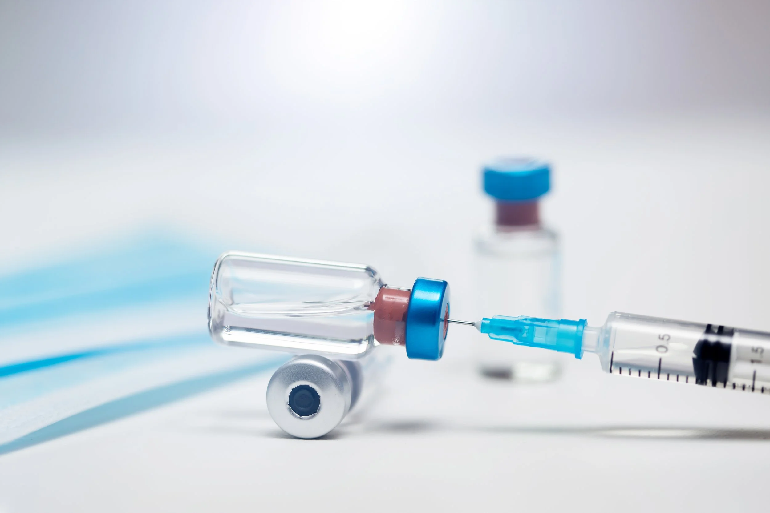 Small glass vials with blue caps beside a syringe drawing liquid from one of them, on a white surface.