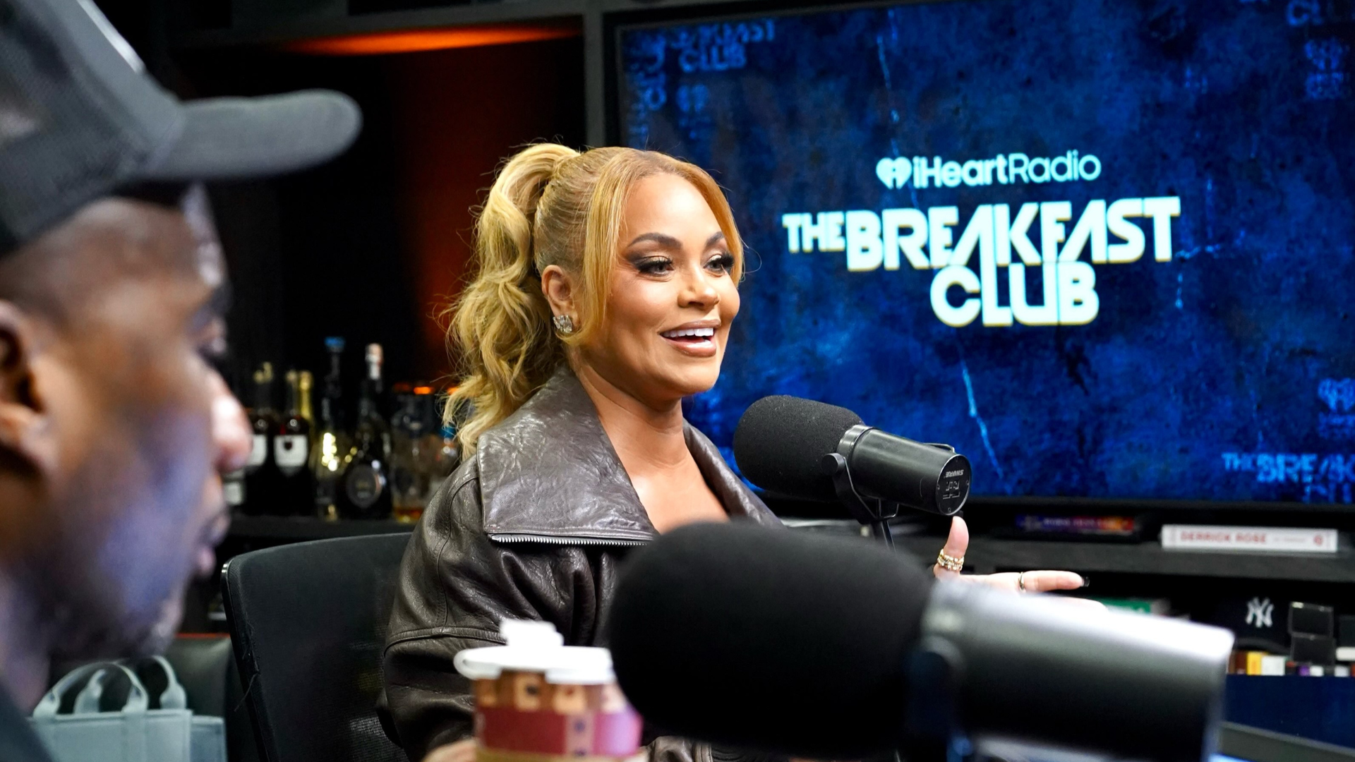 Gizelle Bryant, in a brown leather jacket with her hair in a high ponytail, smiles while speaking into a microphone in a radio studio, with a “The Breakfast Club” screen behind her and co-host Charlamagne Tha God in the foreground