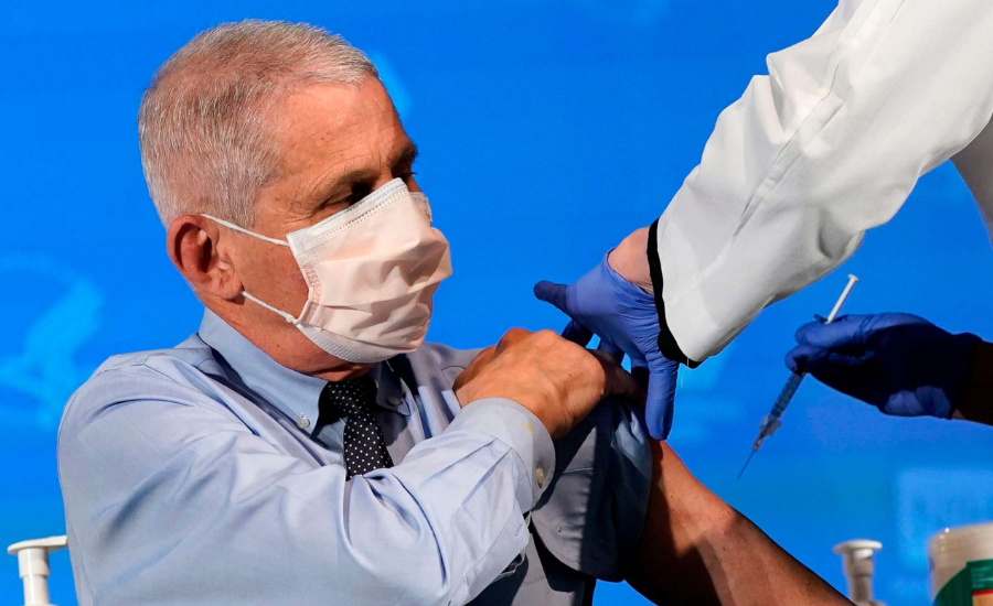 Anthony Fauci, wearing a face mask and dress shirt, rolls up his sleeve as a gloved healthcare worker prepares to administer a vaccine injection.