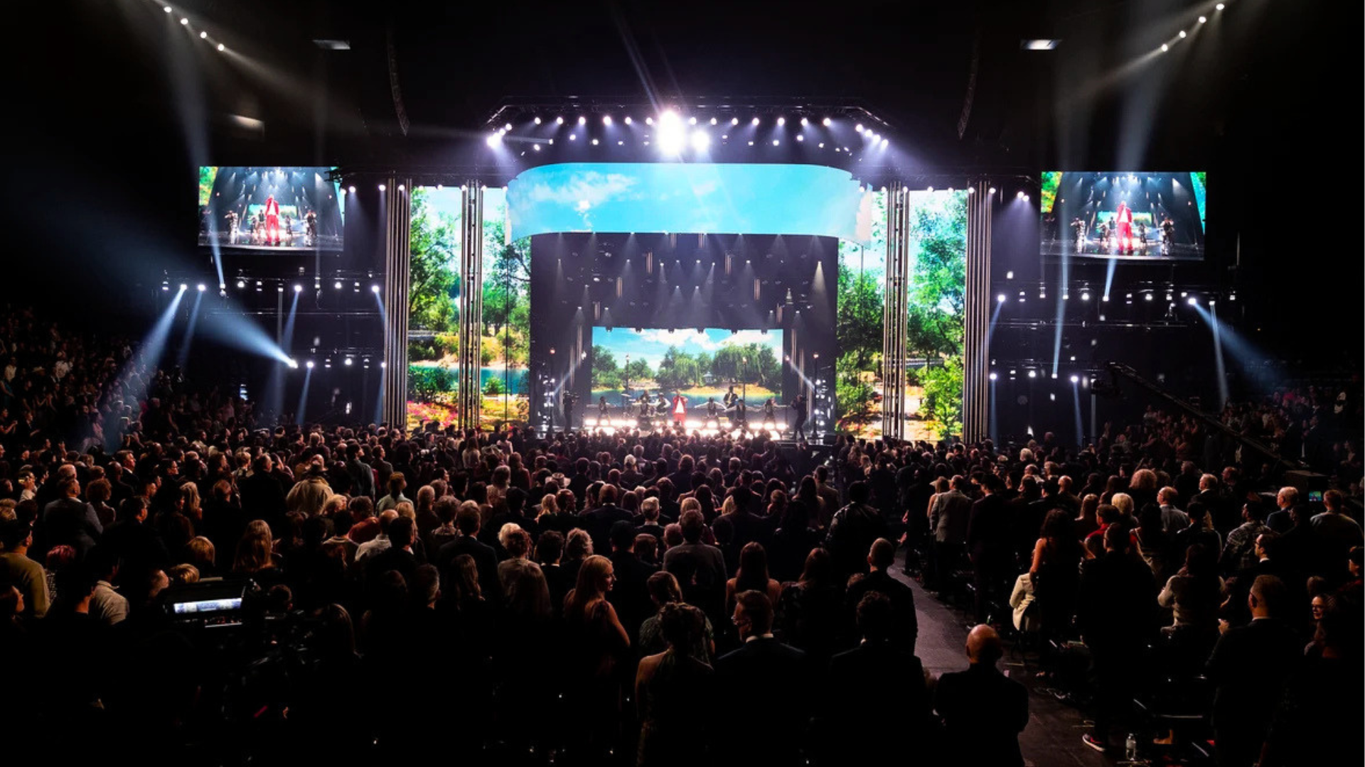 A packed arena audience faces a brightly lit stage where performers stand before large video screens showing a sunny park scene, with spotlights sweeping overhead