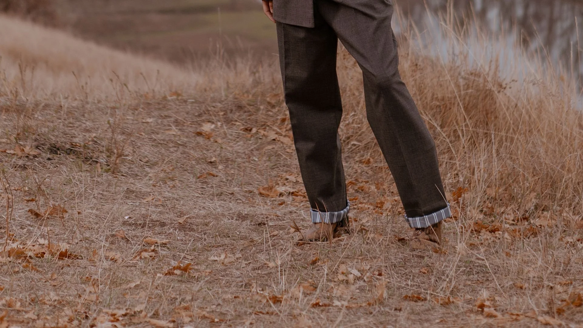 A person in gray trousers and brown boots stands on a path of dry grass and fallen leaves near the edge of a field.