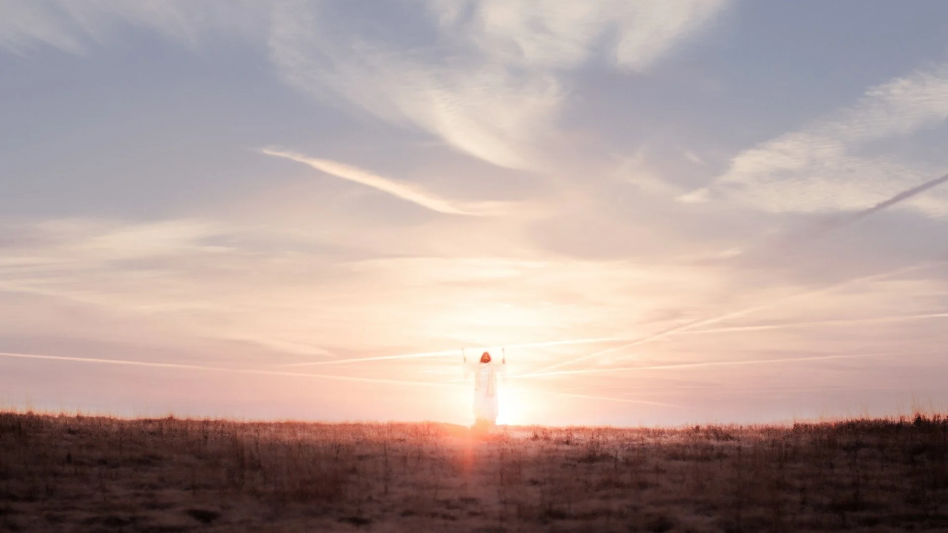 A lone figure in a white robe stands with arms raised in an open field at sunrise beneath a wide, pale sky.