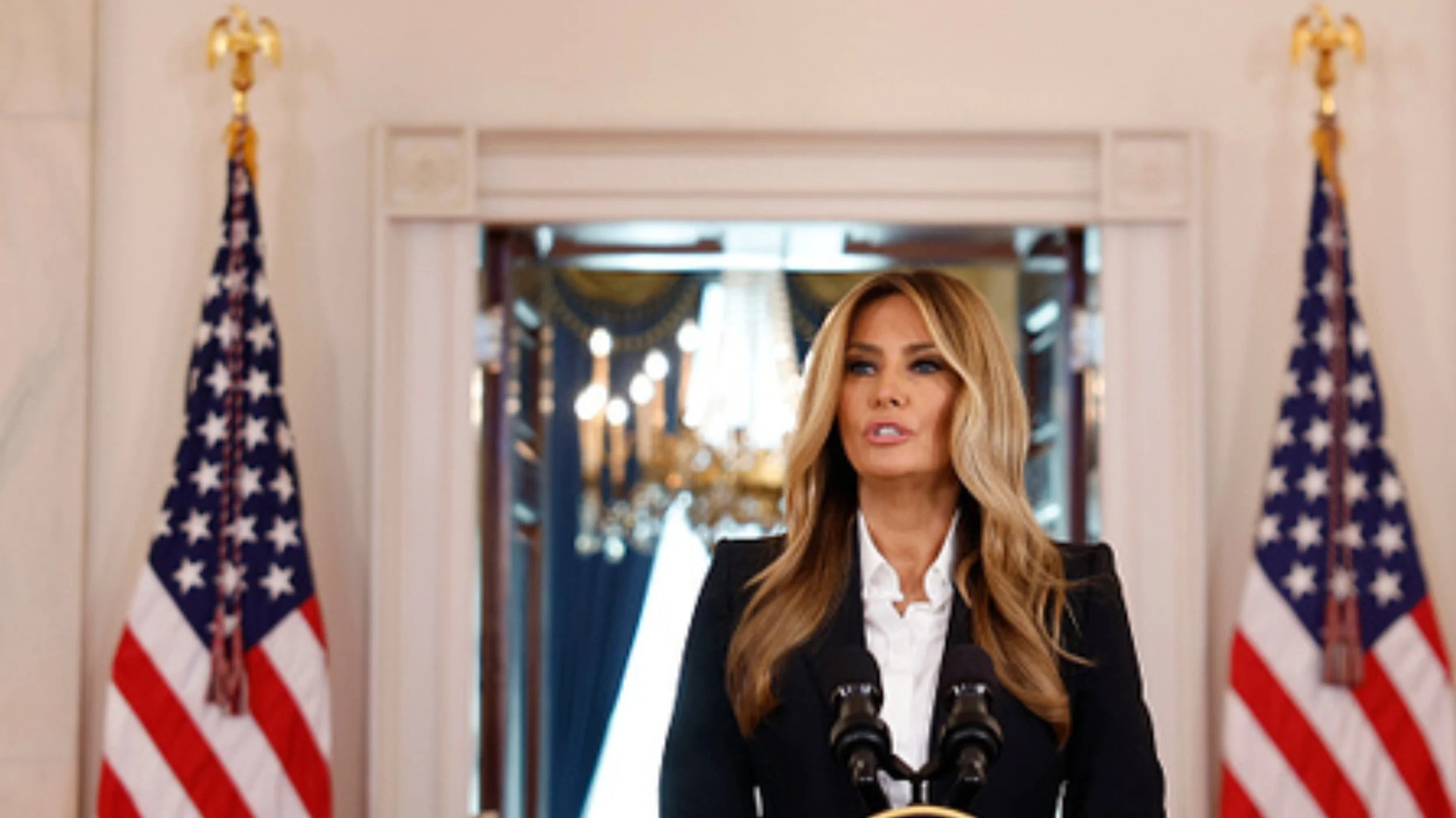 Melania Trump, in a black blazer and white shirt, speaks at a lectern flanked by two American flags in the White House Grand Foyer.