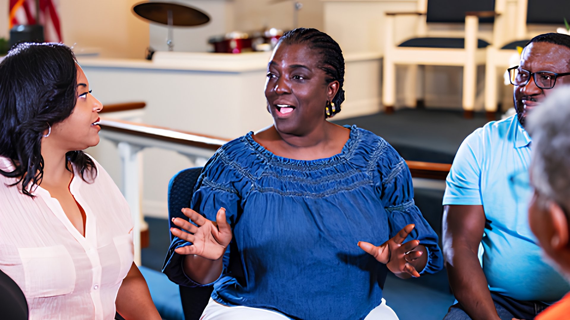 A woman in a blue blouse speaks and gestures as she sits in a circle with other adults inside a church sanctuary, with a pulpit and an American flag in the background.