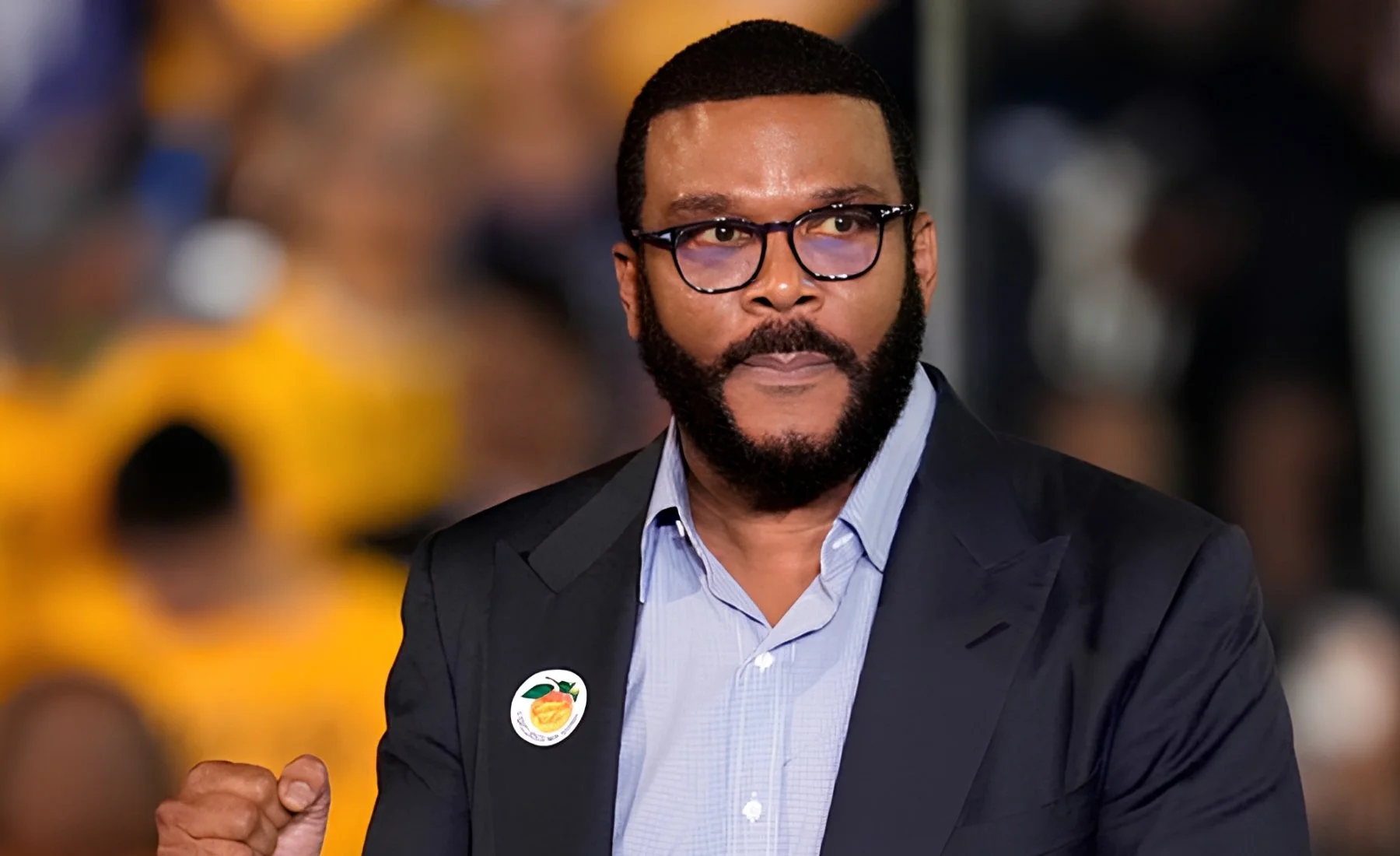 Tyler Perry in glasses and a navy blazer raises a fist in front of a blurred crowd