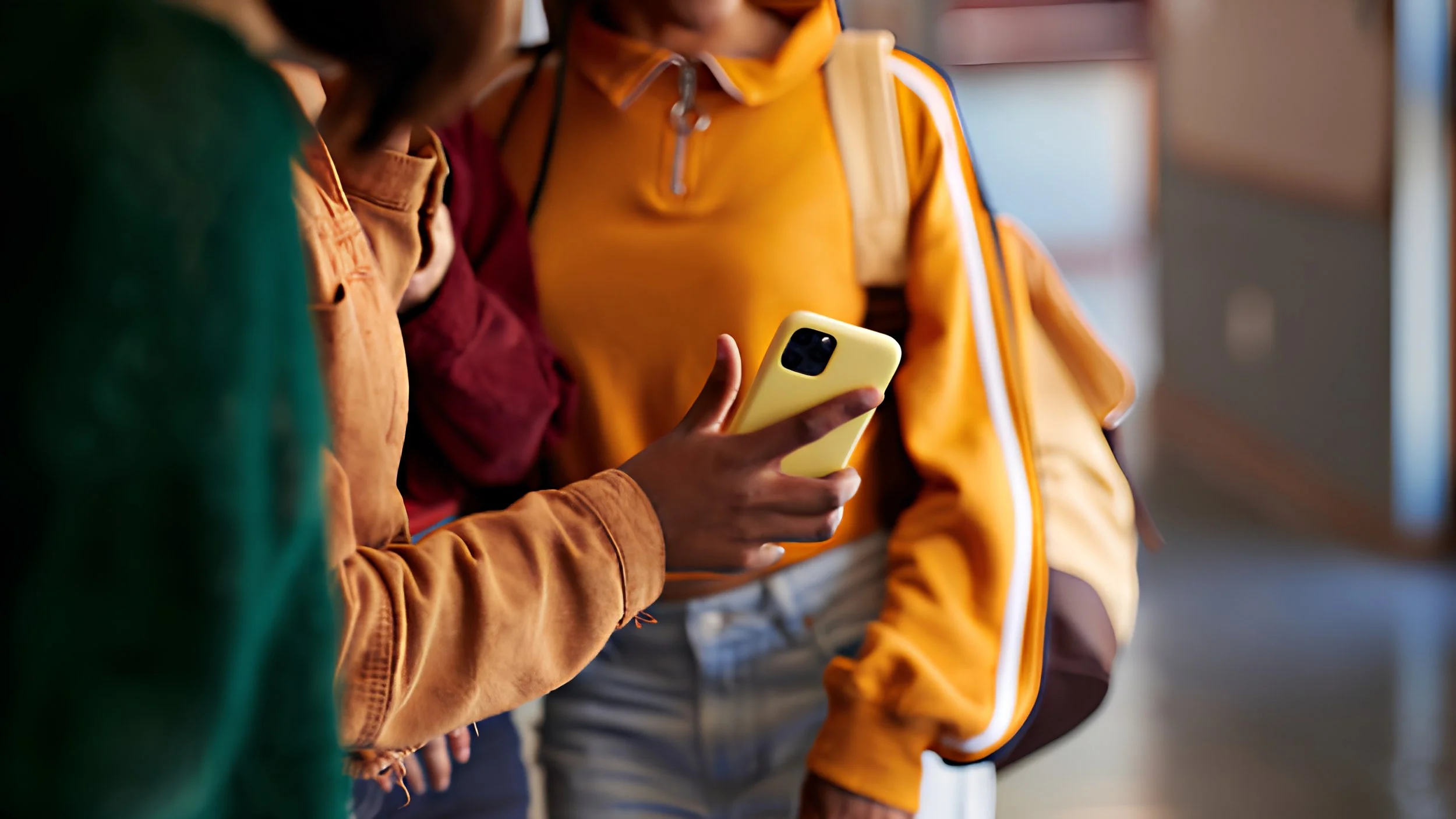 Students in a school hallway, one wearing a backpack and another holding a yellow smartphone.