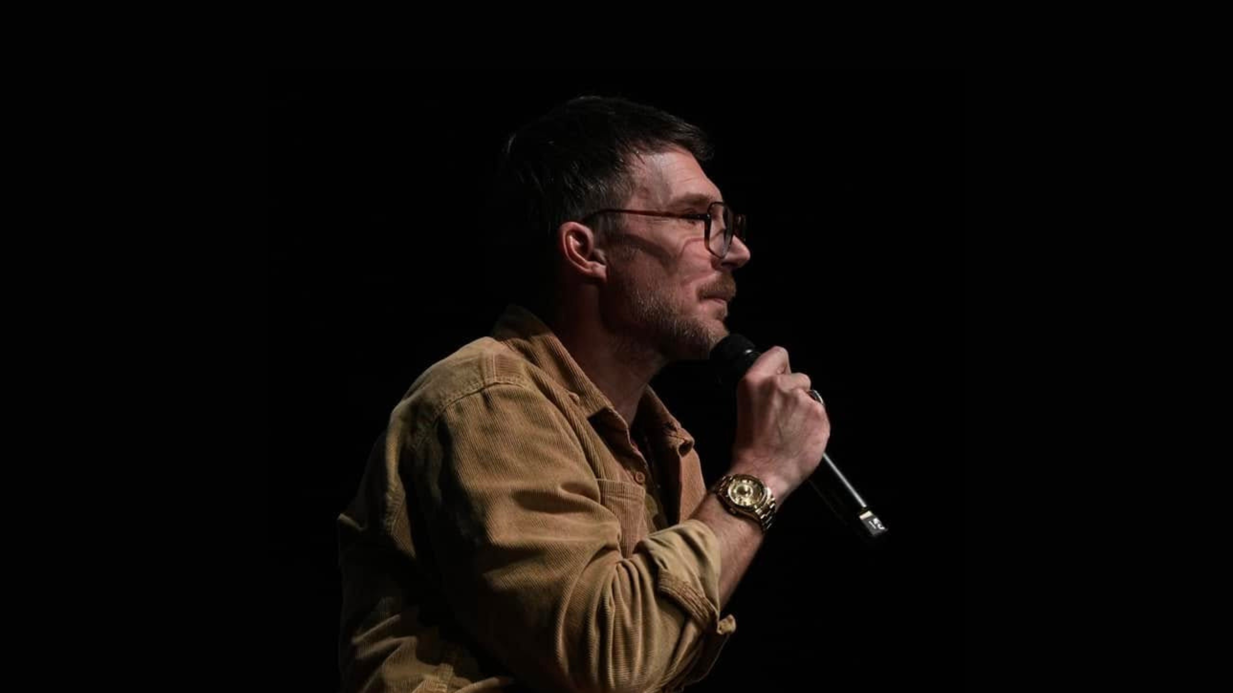 Pastor Judah Smith, wearing glasses and a tan corduroy shirt, holds a microphone while speaking in profile against a dark background.