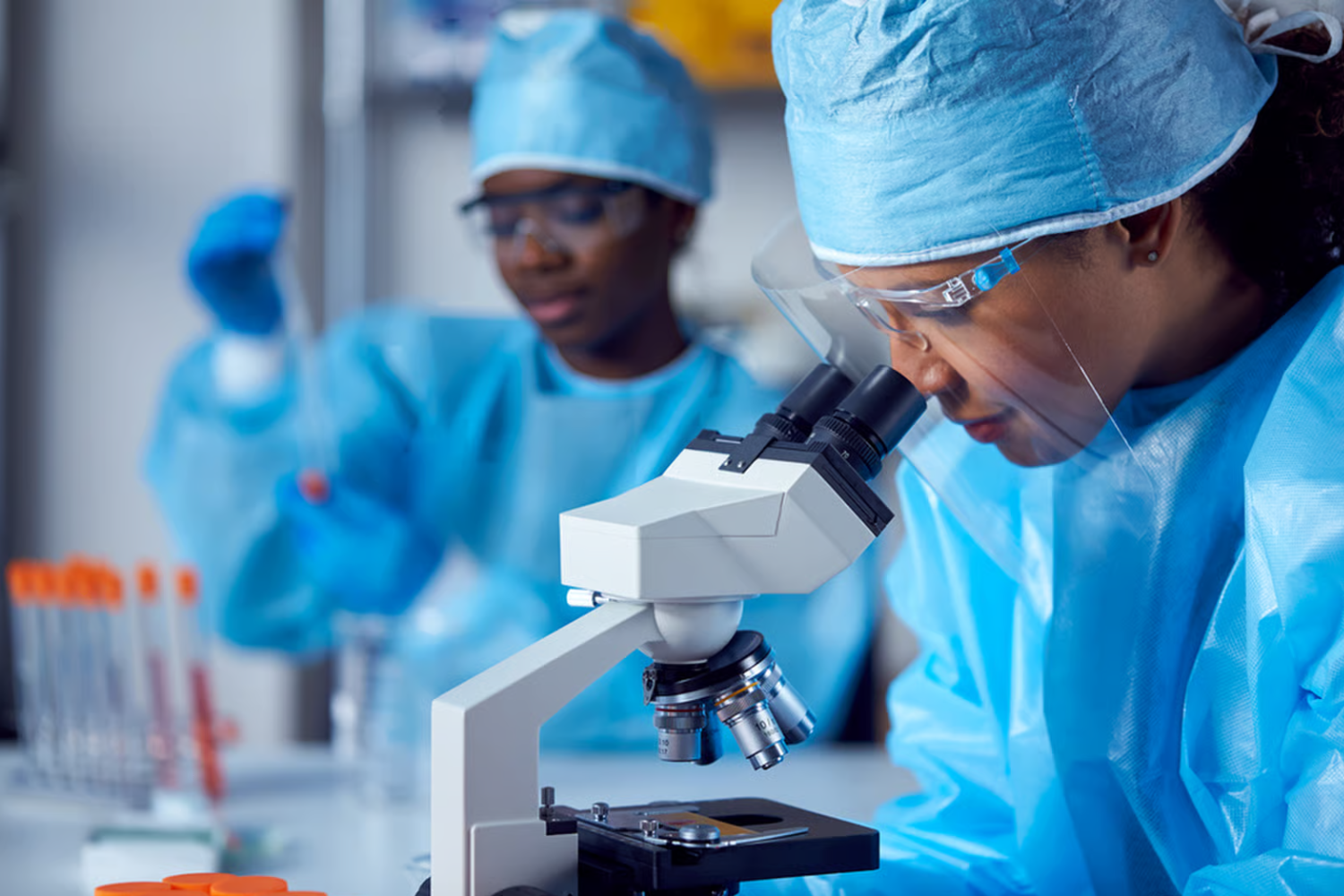 Two scientists in blue protective gowns, caps and safety glasses work in a laboratory. One looks into a microscope while the other handles a sample, with test tubes in the foreground.