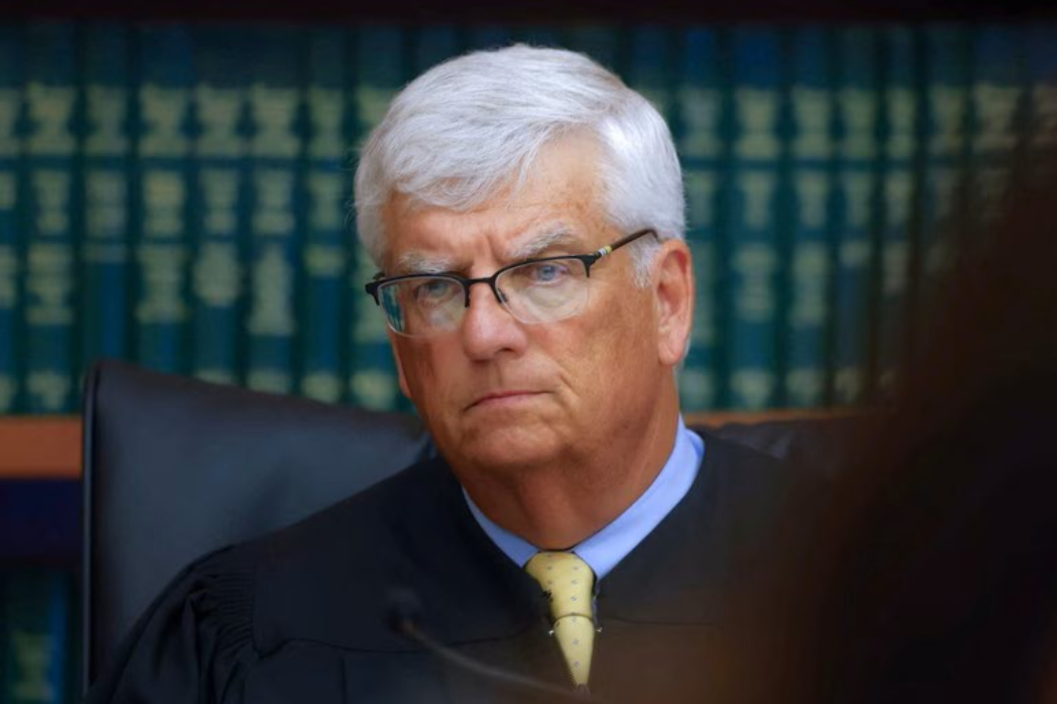 Judge William Sullivan, a man with white hair and glasses, wearing a black robe and a yellow tie, sits on the bench in front of shelves of law books.