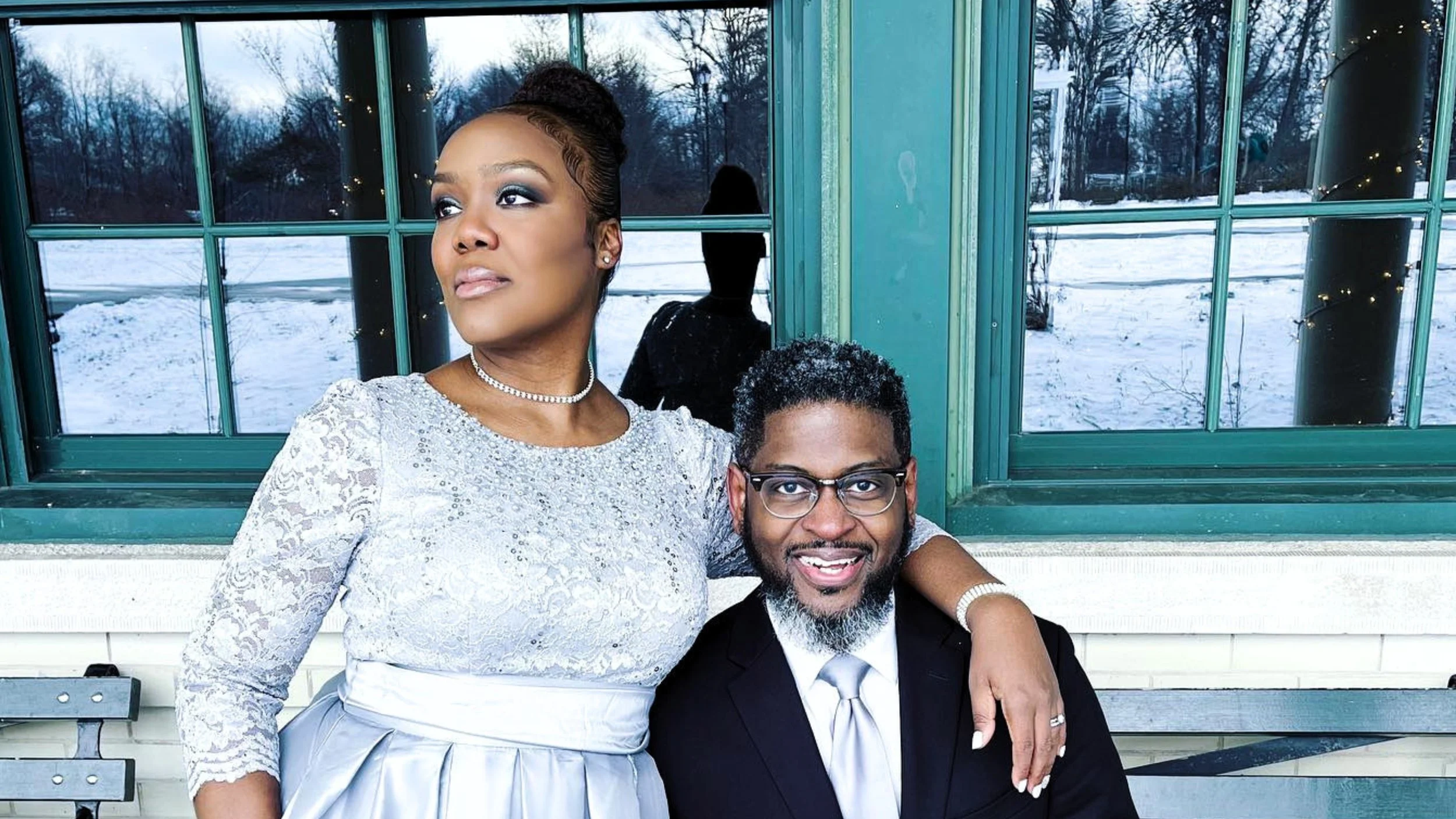 Dianna Hobbs, wearing a silver lace gown, rests her arm around her husband, Kenya Hobbs, who sits smiling in a black suit and silver tie in front of green-framed windows overlooking a snowy park.