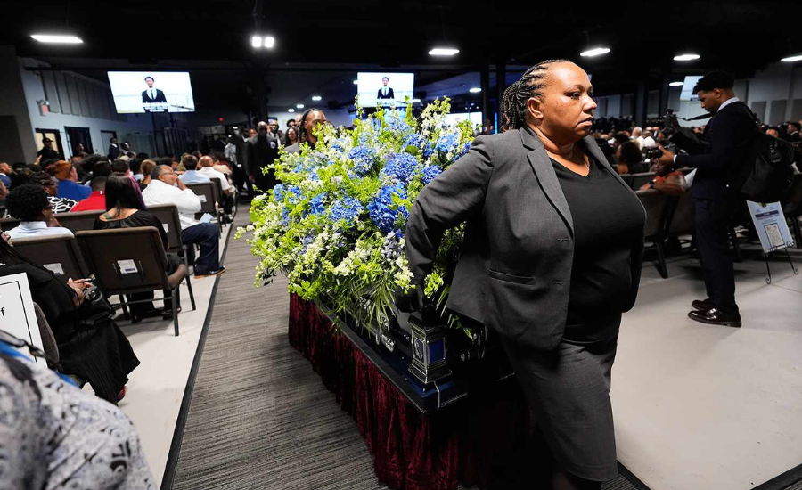A woman stands near a floral memorial display at a funeral service, with mourners seated in the background