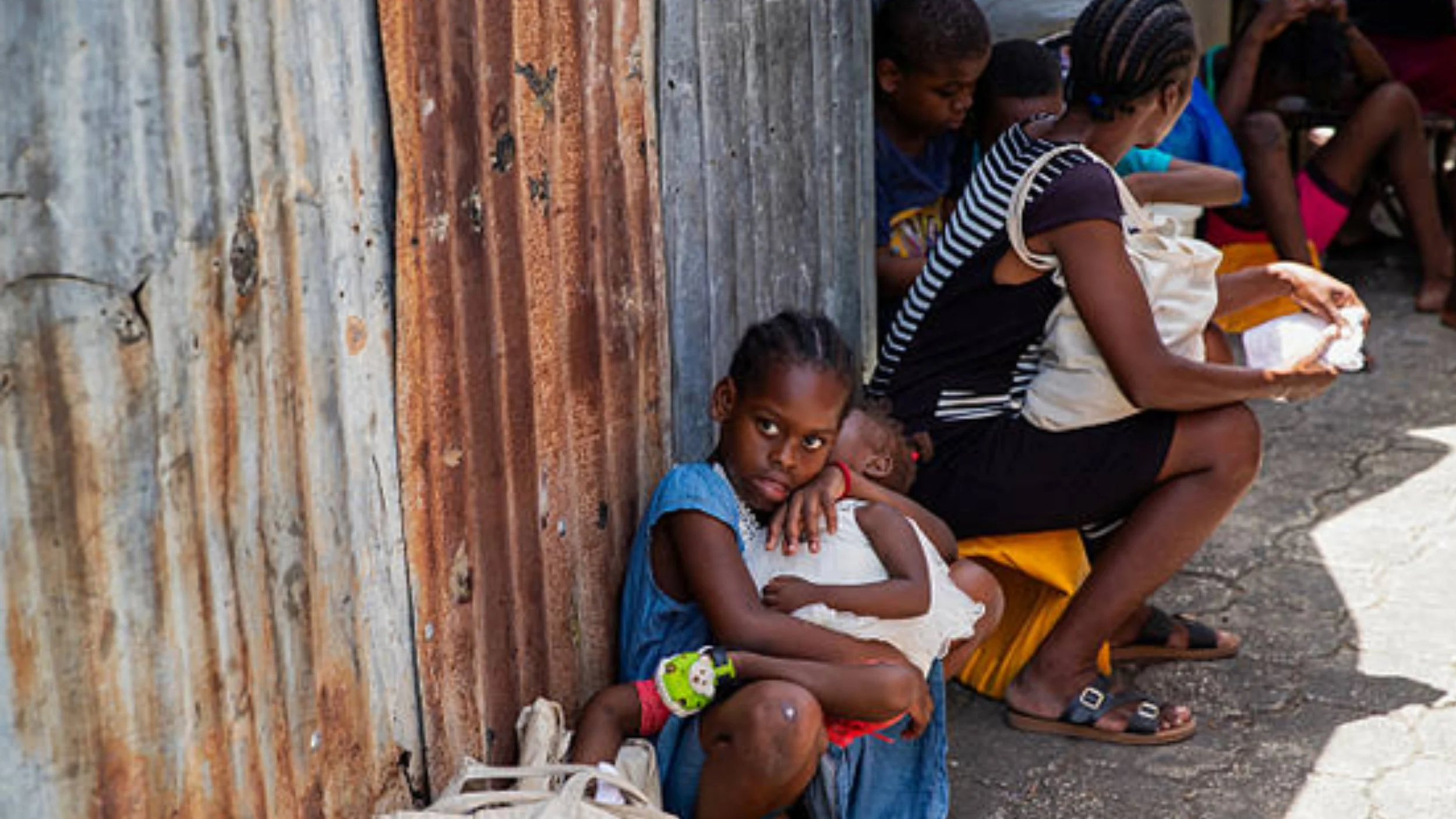 A girl sits against a rusted corrugated metal wall, holding a sleeping toddler in her lap, as a woman and other children sit nearby at a camp.
