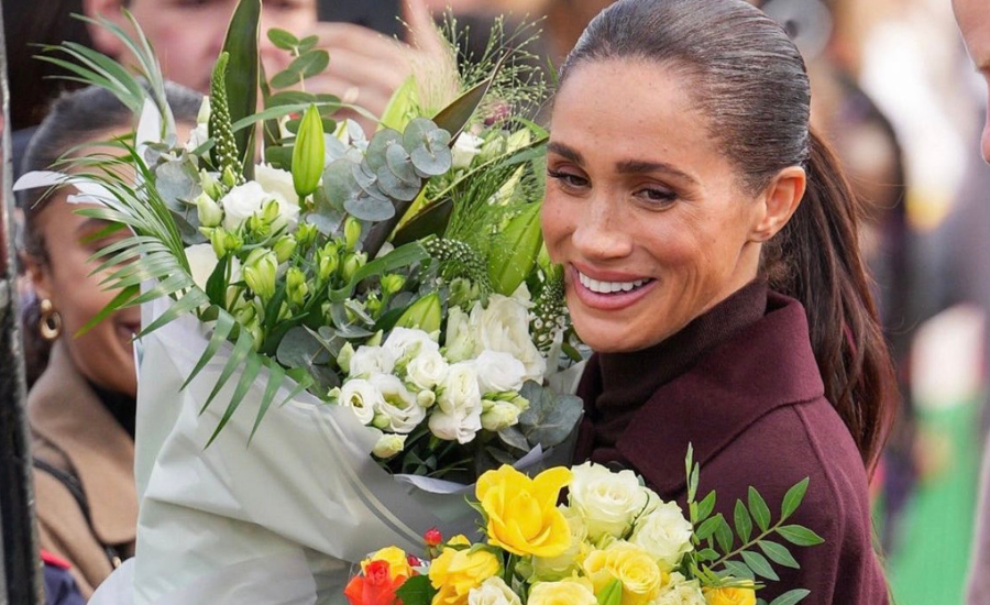 Meghan, in a burgundy coat, smiles while holding a large bouquet of white flowers and a smaller bouquet of yellow and white roses.
