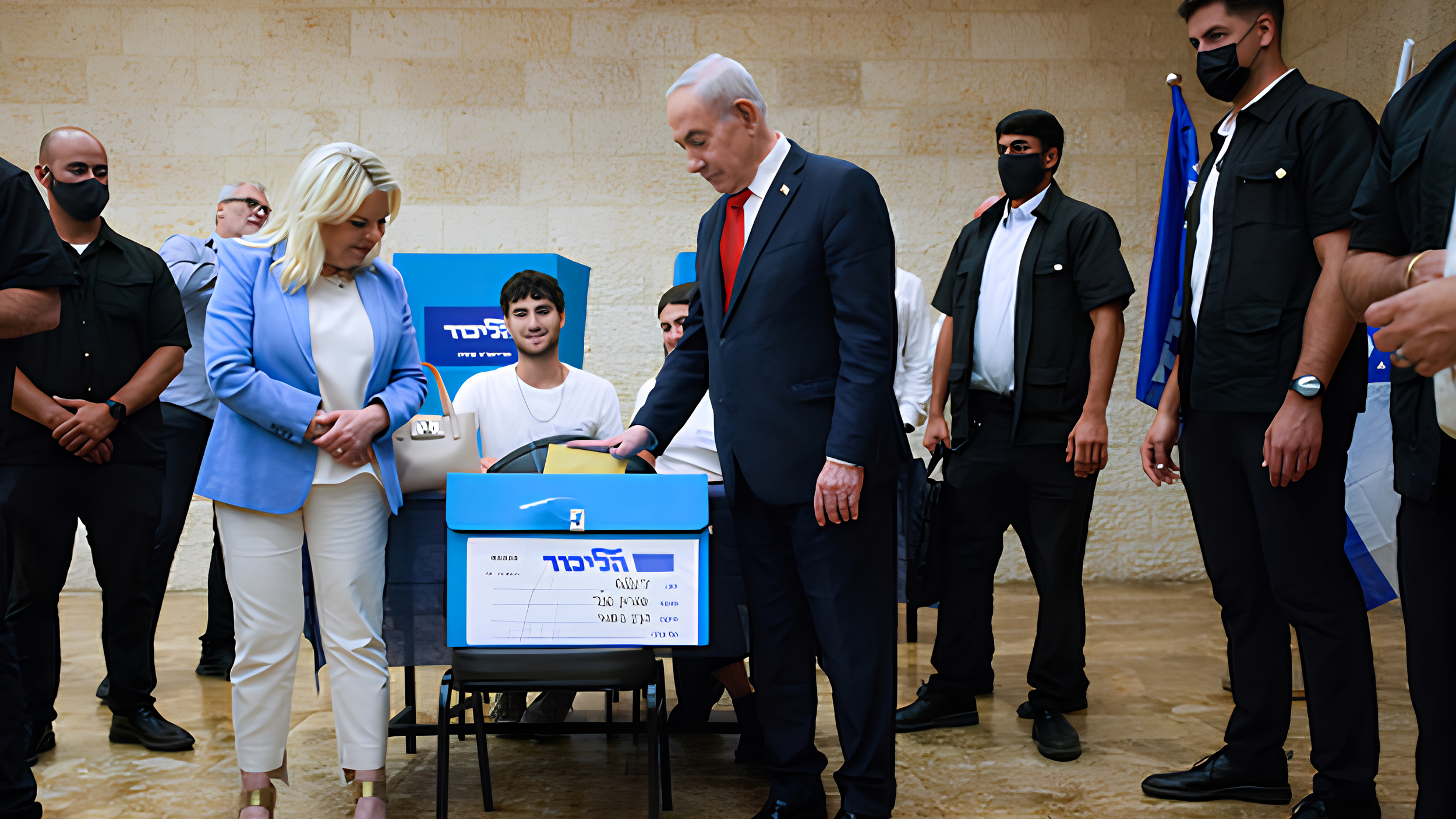 Benjamin Netanyahu drops a ballot into a blue Likud ballot box as his wife, Sara, stands beside him and security officers look on, in a stone-walled room in Jerusalem.