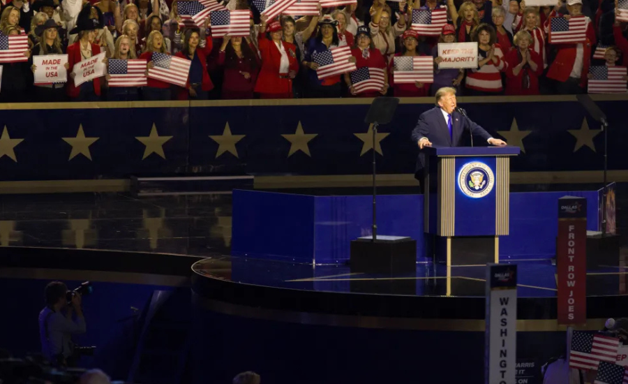 President Donald Trump speaks at a podium during the 2026 Republican Convention in Dallas as supporters behind him wave American flags and hold signs.