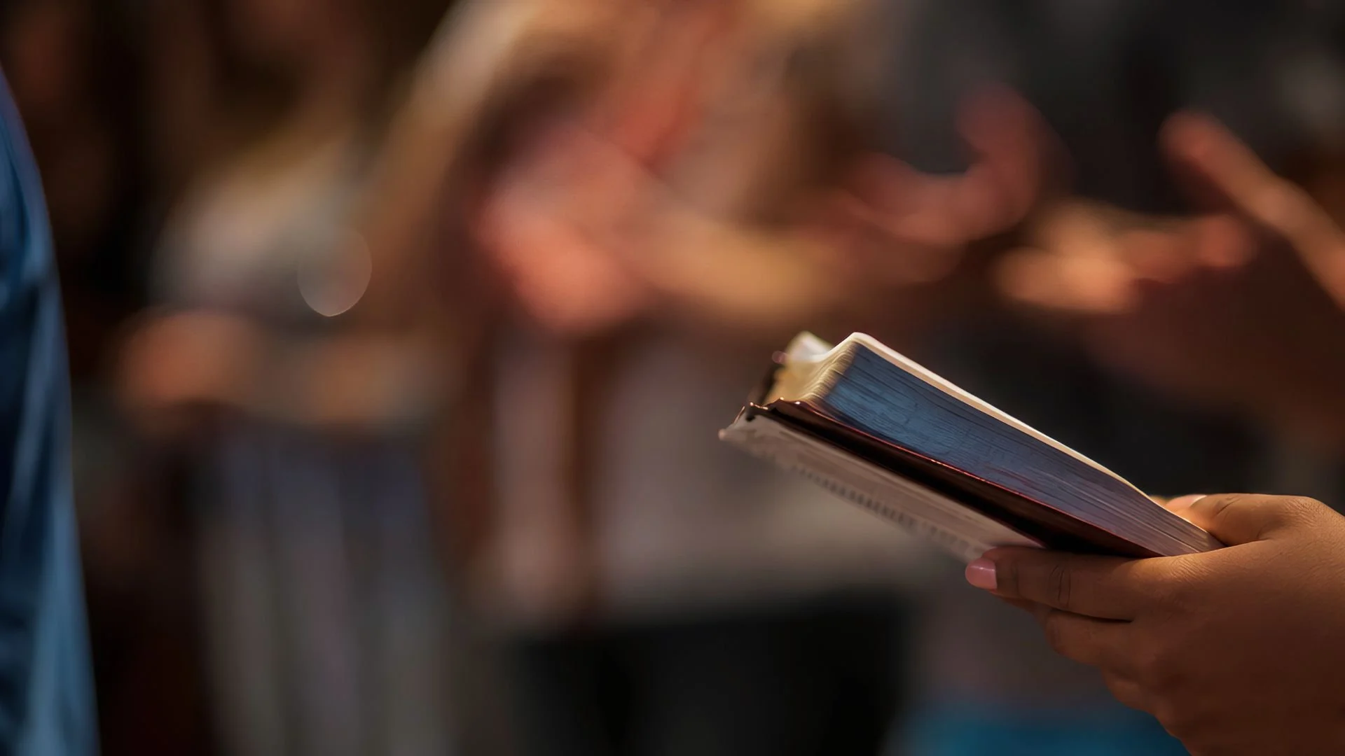 A hand holding a worn Bible in the foreground as worshipers raise their hands in the blurred background.