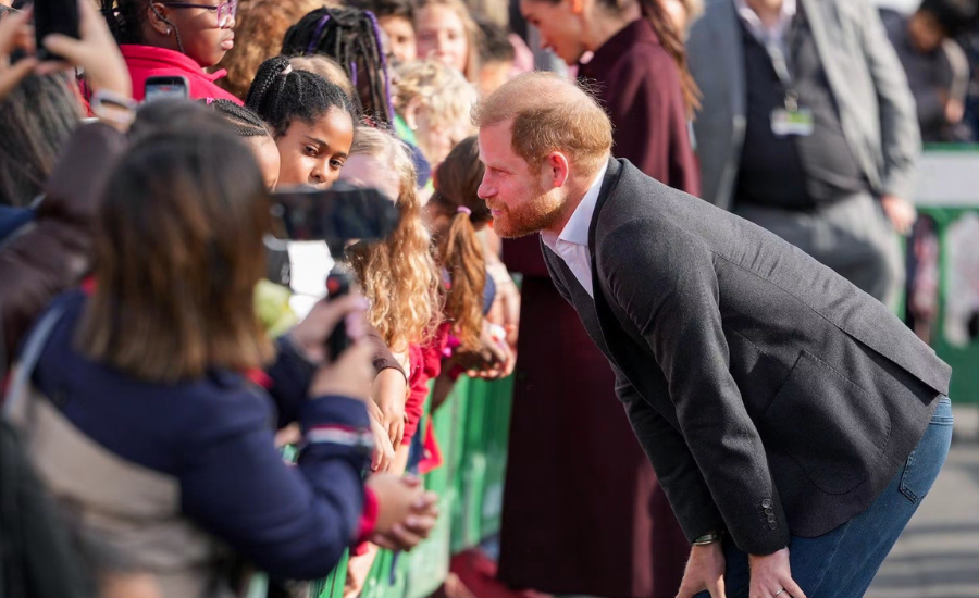 Prince Harry bends forward to speak with a group of young girls gathered behind a green barrier, one holding up a camera.