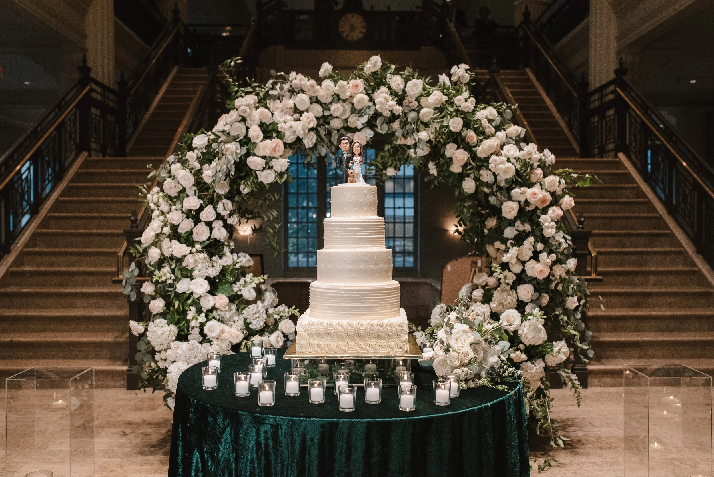 A towering circular floral arch overflowing with ivory blooms and cascading greenery frames the wedding cake at The Corinthian Houston.