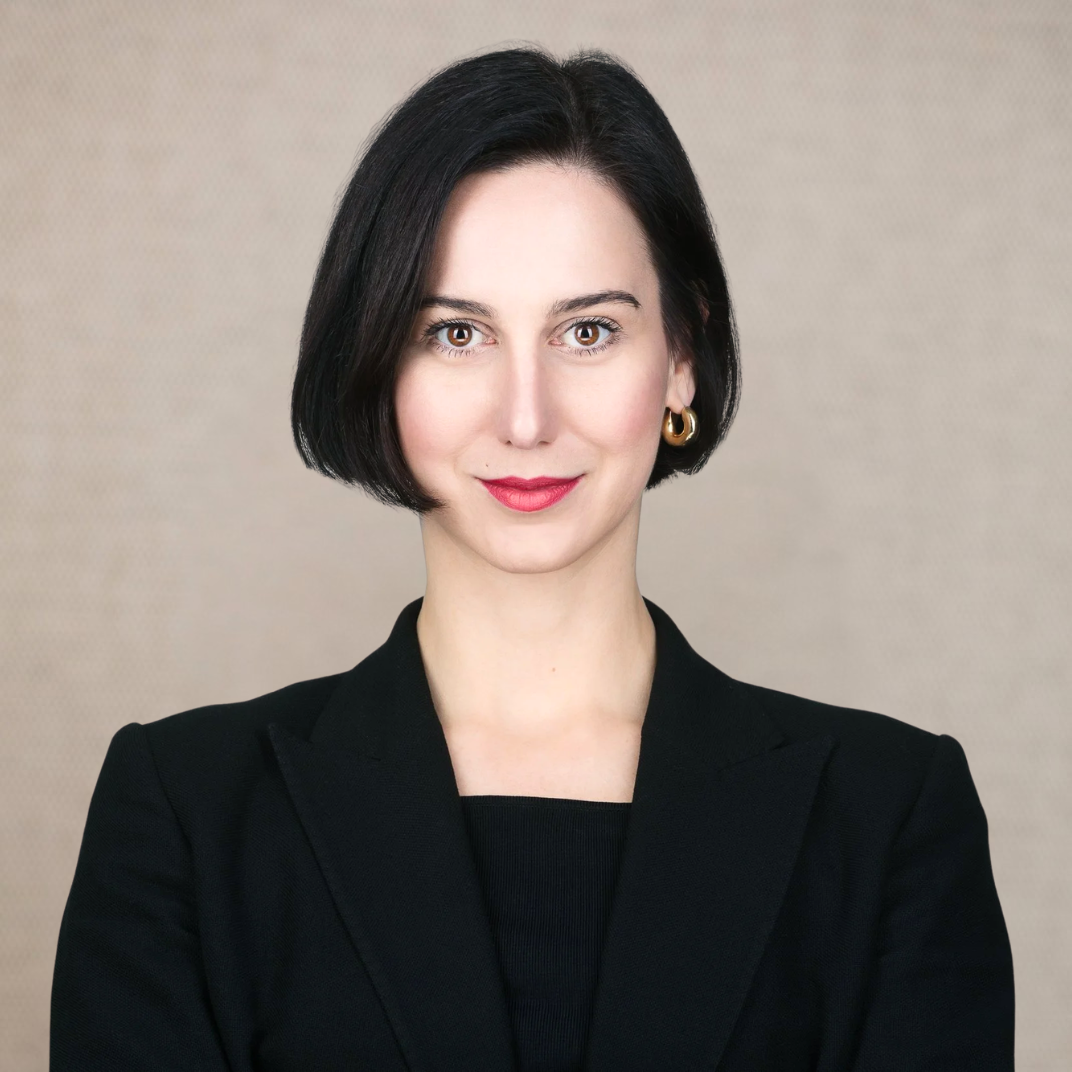A woman with short black hair, red lipstick, wearing gold hoop earrings and a black blazer against a plain white background.