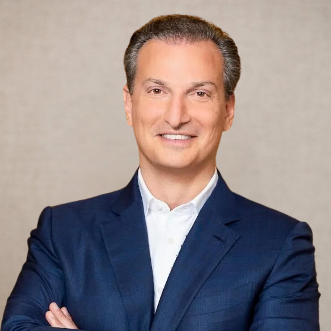 A man in a navy suit with a white shirt, smiling with arms crossed, standing in an office setting.