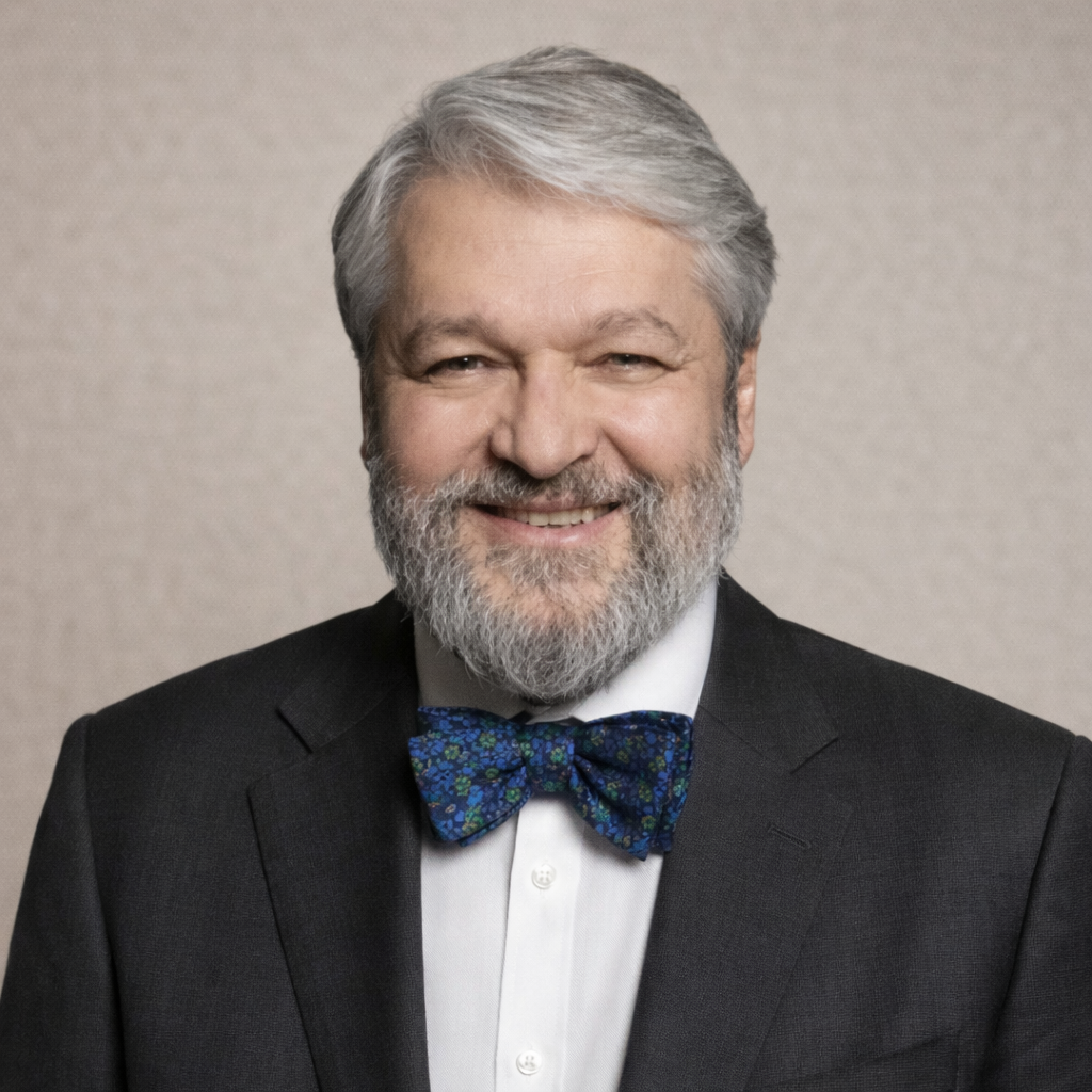A smiling older man with gray hair and a beard, wearing a dark suit, light shirt, and bow tie.