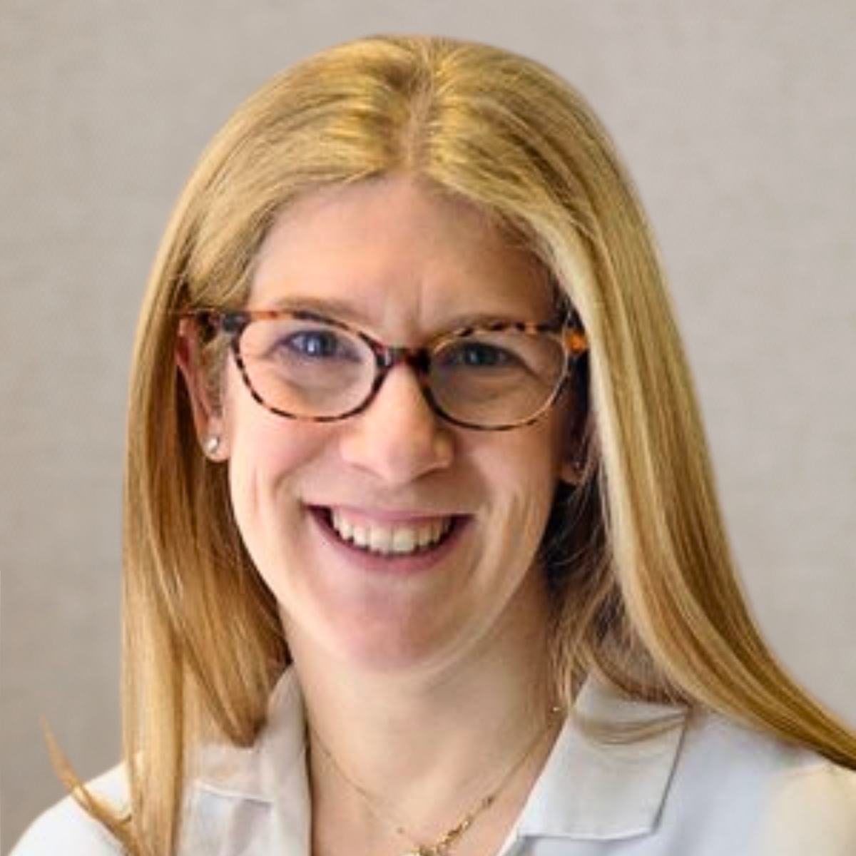 Portrait of a woman with blonde hair, glasses, and a white medical coat with green embroidery, smiling in a clinical setting.