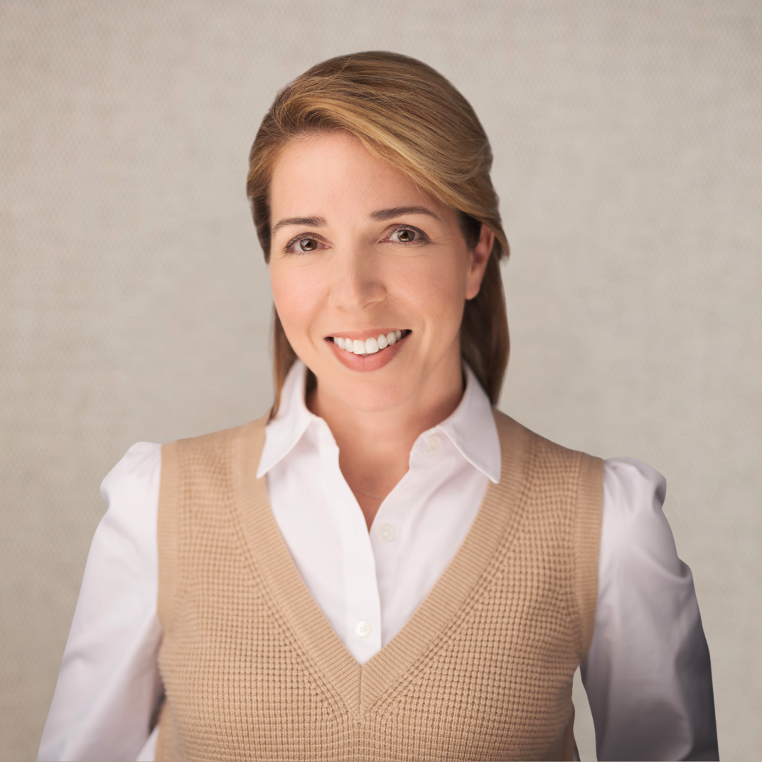 A woman with short, light brown hair smiling, wearing a white blouse and a beige sweater vest.