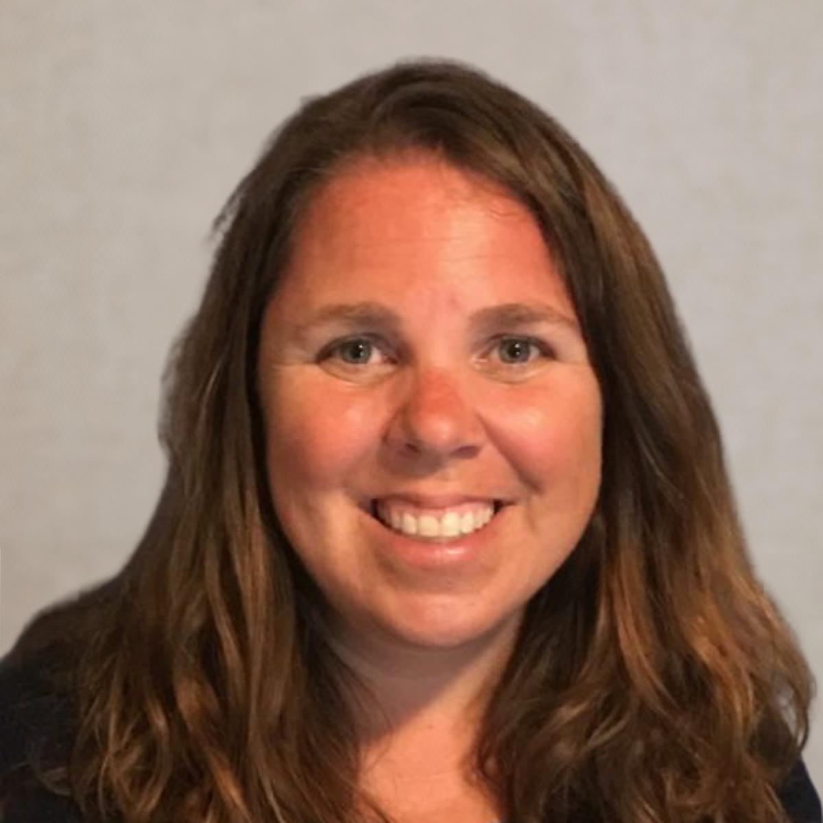A woman with long, wavy brown hair smiling at the camera, wearing a dark blazer over a blue patterned top.