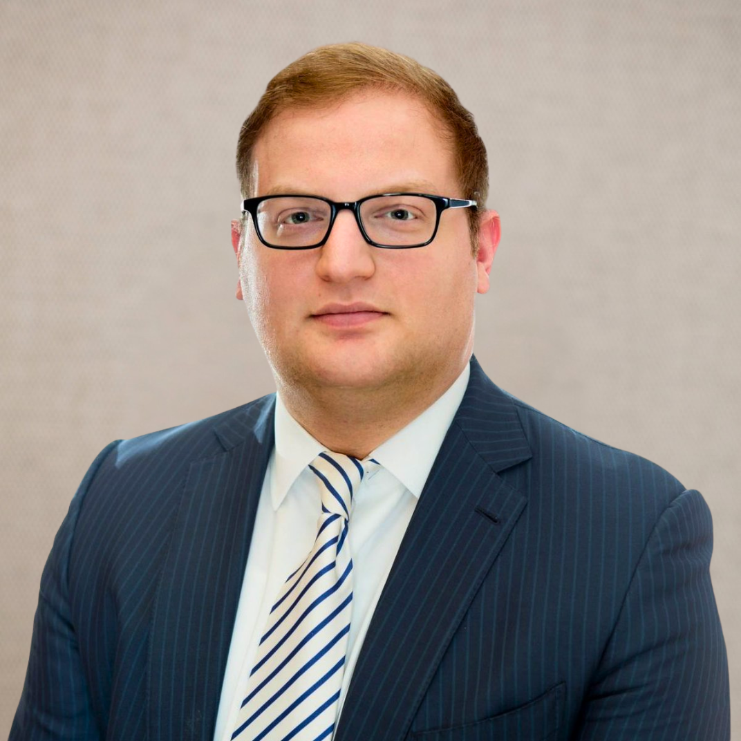 A professional headshot of a man with short red hair, wearing glasses, a dark blue pinstripe suit, white shirt, and striped tie against a neutral background.