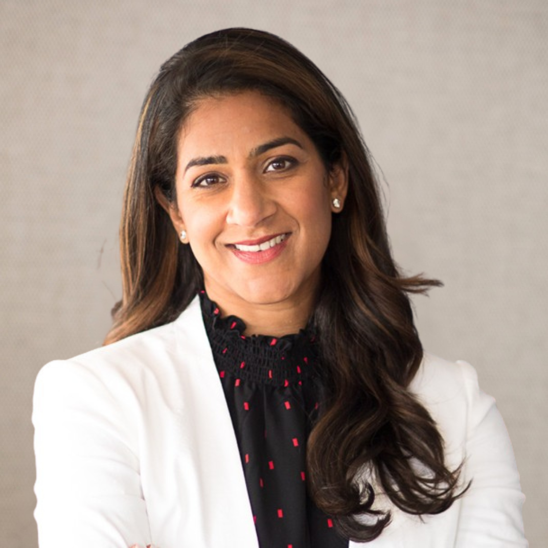 A woman with long dark wavy hair and earrings, wearing a black top and colorful beaded necklace, smiling against a plain light background.