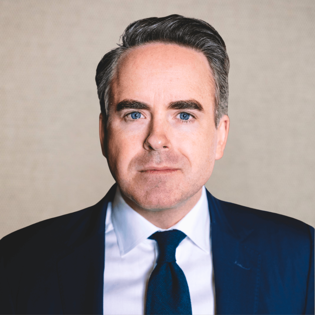 Professional man with light skin, brown hair, and blue eyes smiling, wearing a dark suit and tie, in an office setting.