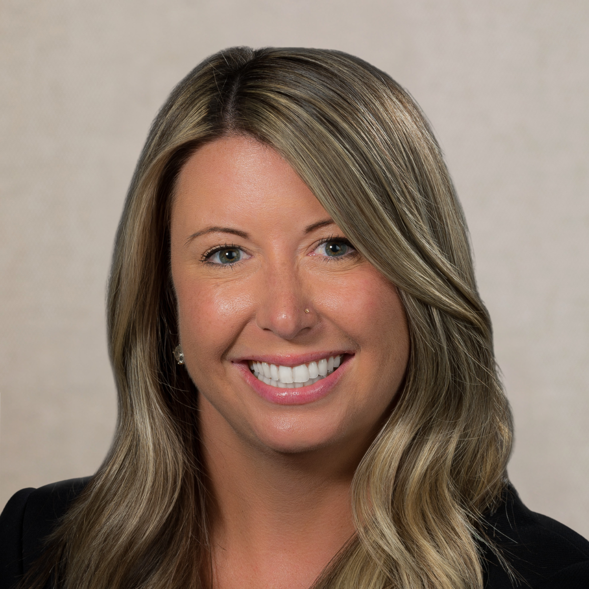 Portrait of a woman with long wavy blonde hair, wearing a black blazer and a patterned blouse, smiling against a gray background.