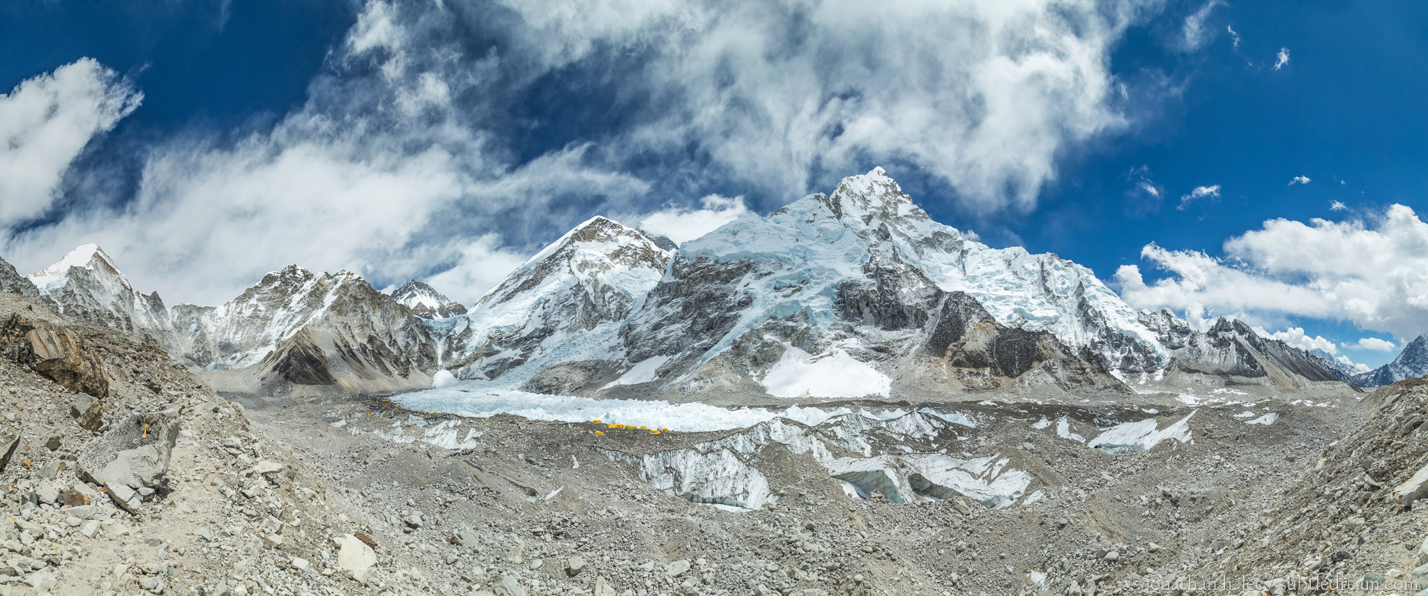 Everest basecamp (yellow/orange tents), Khumbu Glacier (behind basecamp), and Khumbu Icefall (above glacier). Nuptse in the background 7,861 m (25,791 ft) in the center-right. Everest slightly hidden behind the clouds in the middle-back.