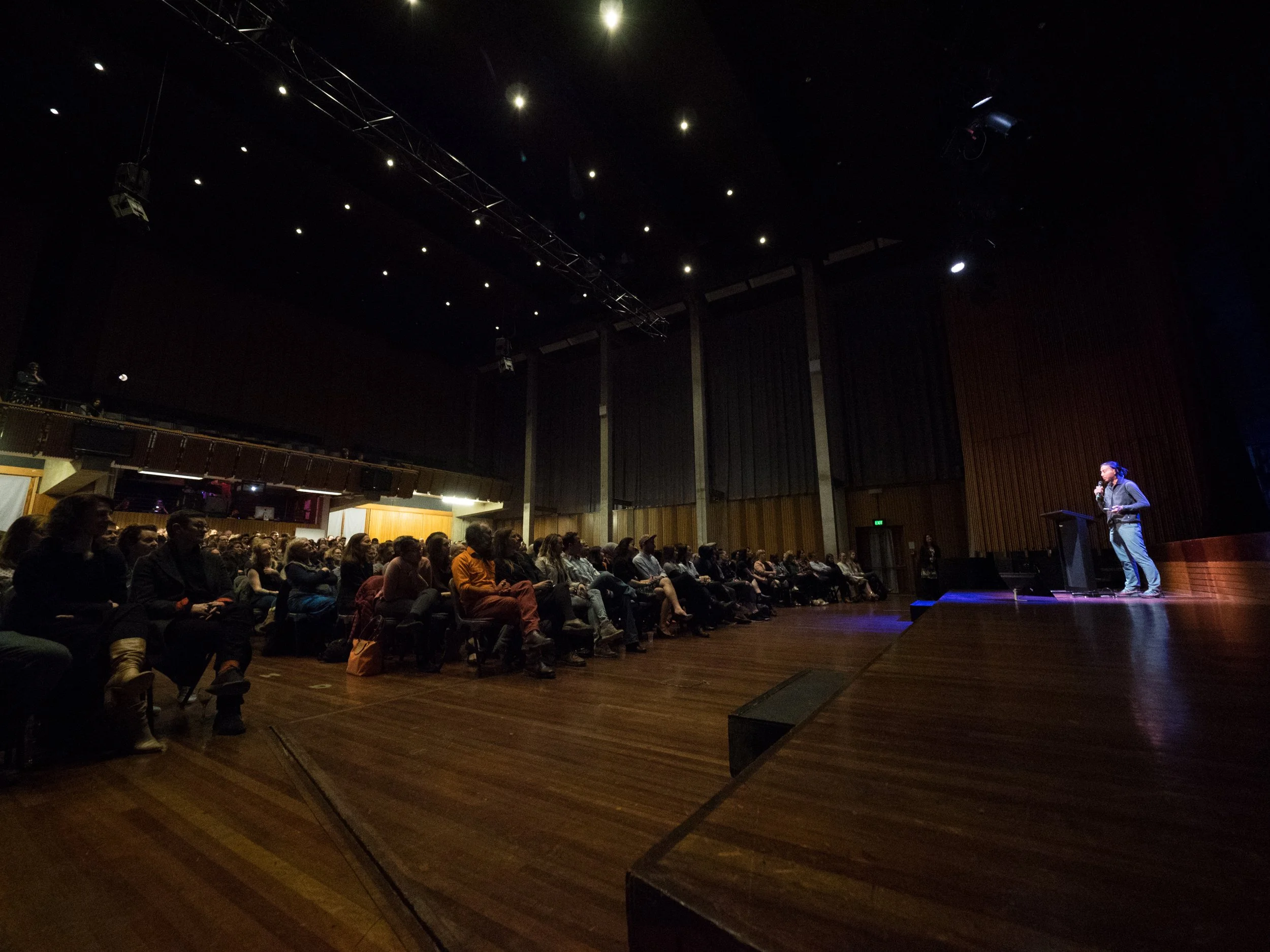 An indoor auditorium with a large audience seated and a woman speaking at a podium on stage. The ceiling has several small lights and stage equipment.