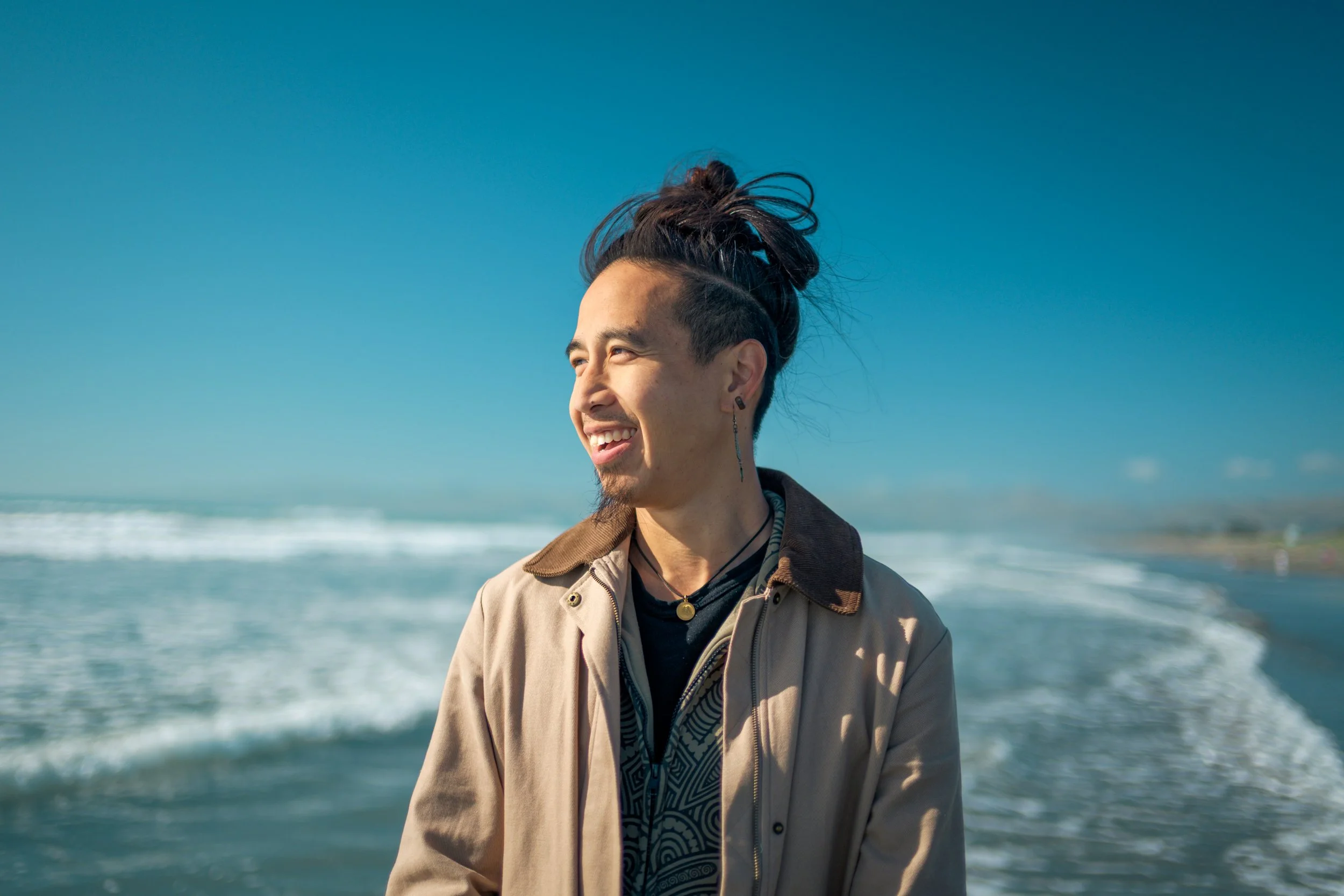 A young man with dark hair and piercings smiling on a beach with waves in the background.