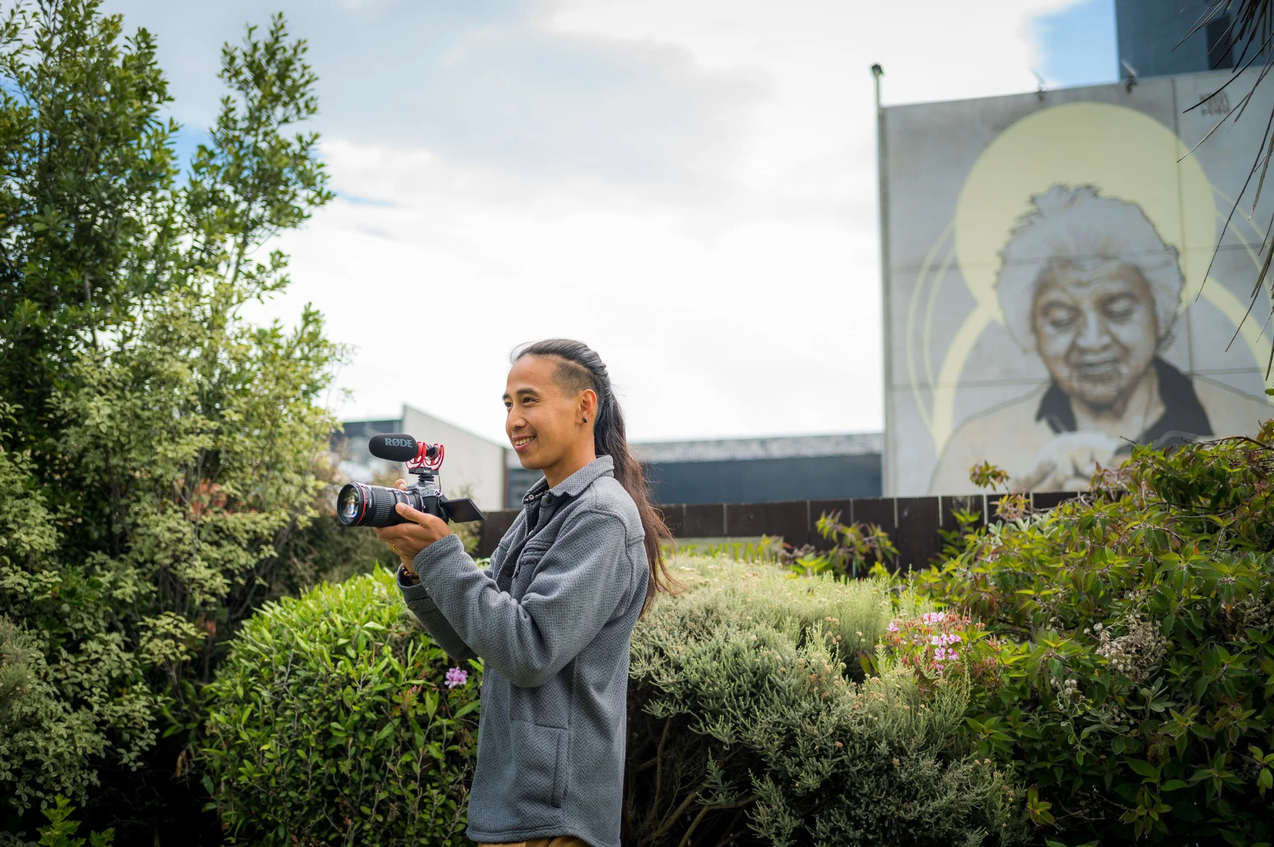 A young man with long hair and a gray blazer holding a camera with a microphone attachment, standing outdoors in a garden area with green shrubs and a mural of a smiling person on a building wall in the background.