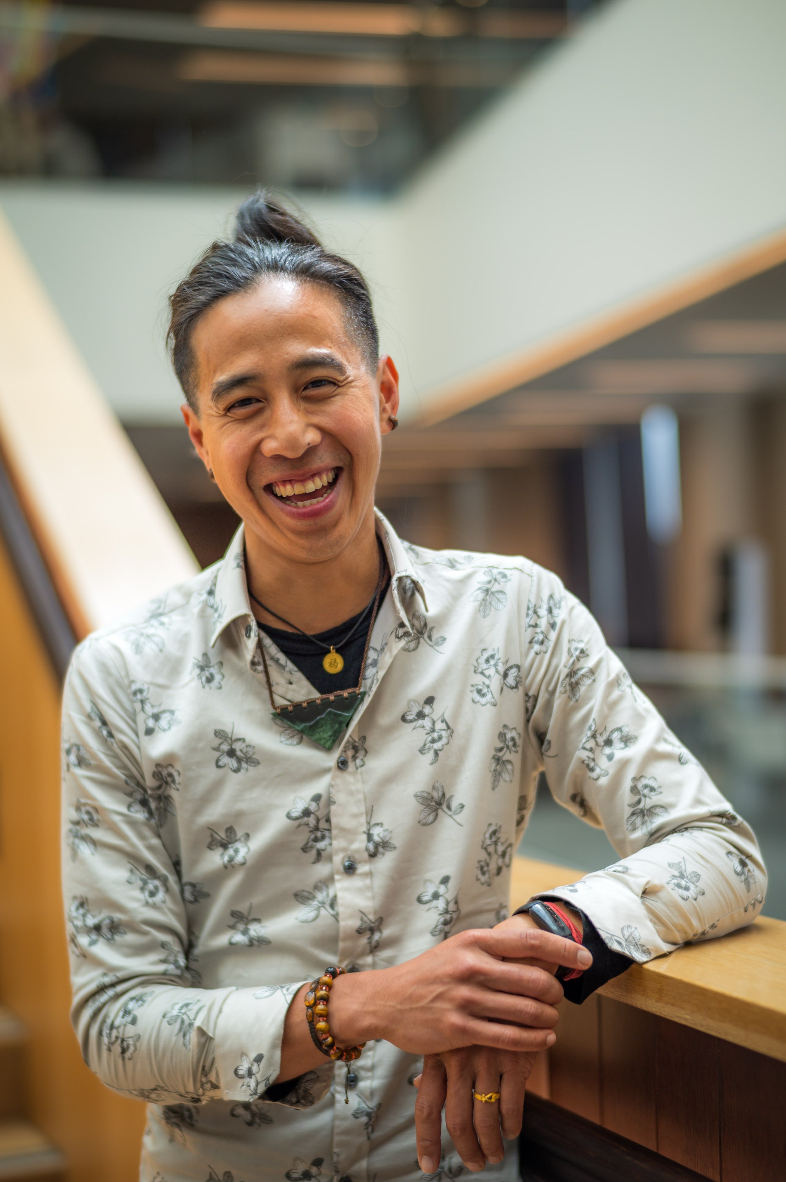 A smiling man with dark hair pulled back, wearing a floral shirt and accessories, sitting at a wooden table in a contemporary indoor setting.
