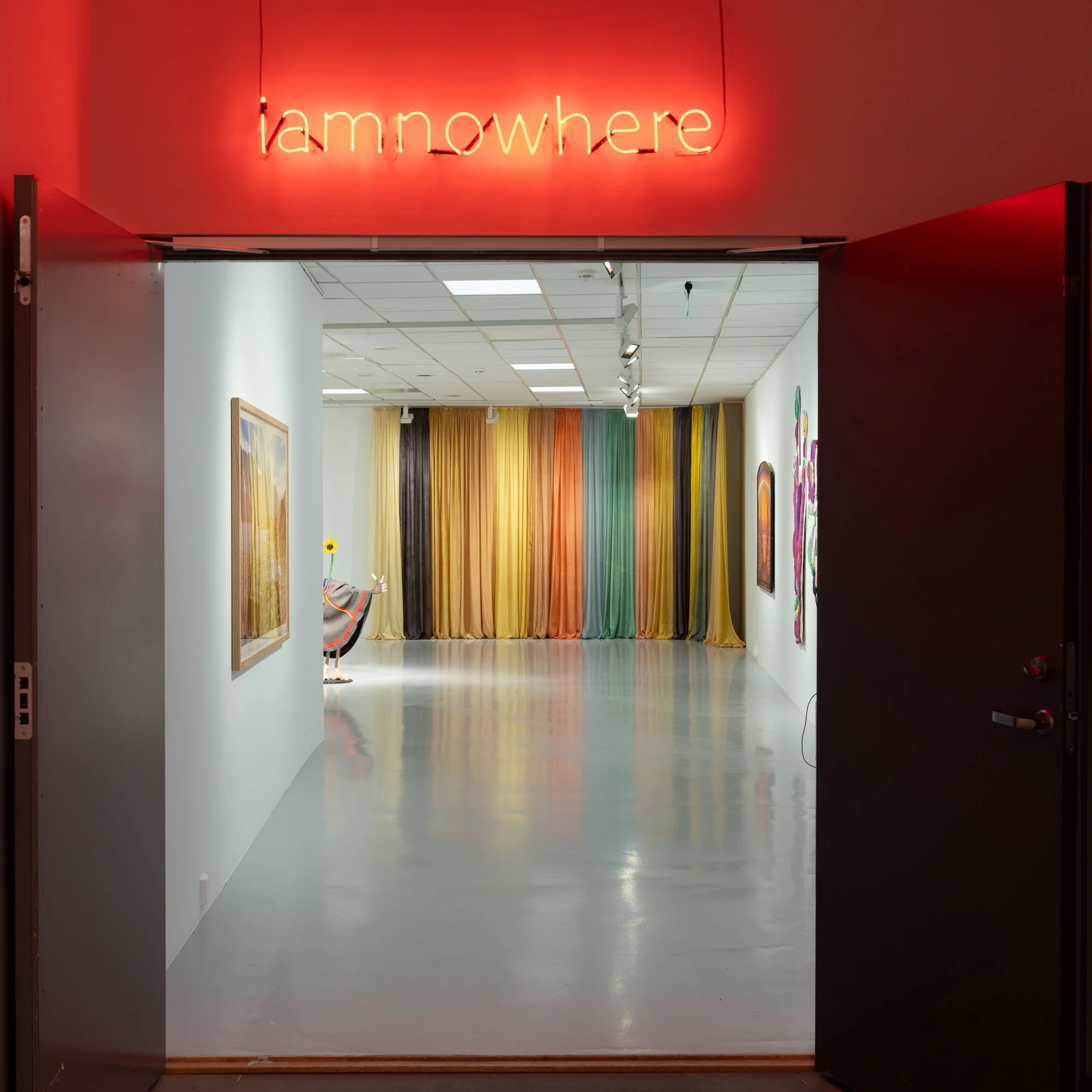   iamnowhere  (2025) av Ina Otzko, lysskilt montert over inngangspartiet til utstillingsrommet. På veggen i bakgrunnen henger Hilde Hauan Johnsens tekstilverk  Fargeprisme fra natur IV   (2022–2024). Foto: Kjell Ove Storvik / Nordnorsk kunstnersenter