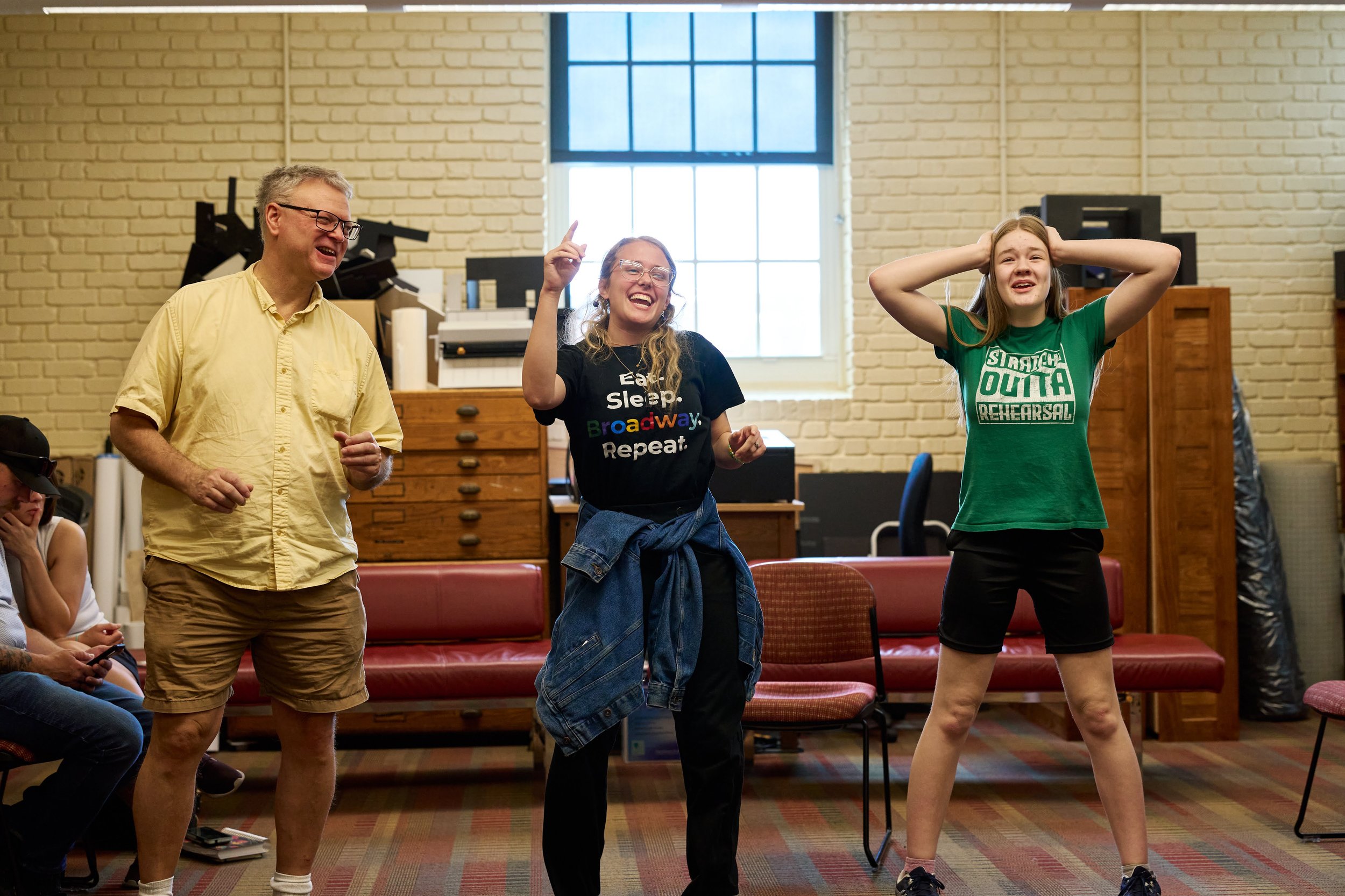 3 adults stand next to each other horizontally: a man wearing a yellow shirt laughing, a woman with glasses and blonde hair pointing up and laughing and a woman wearing a green shirt covering her ears and laughing. They are in the middle of an improv