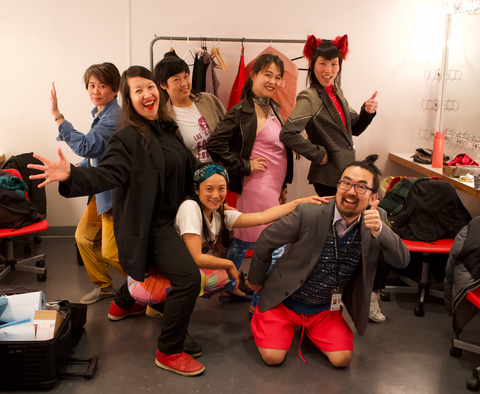 Left to right, members of FXR Shirley Tse, Amanda Ross-Ho, Patty Chang, Anna Sew Hoy, Amy Yao, Pearl  C. Hsiung, Ei Arakawa-Nash posing together giving thumbs up and jazz hands in a theatre backstage dressing room